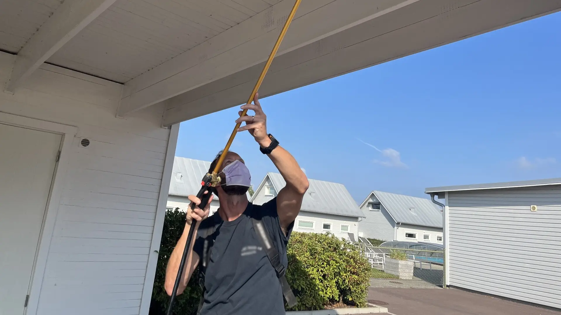 Anders Olsson, spindelsanerare på Öland, använder en lång saneringsstav för att ta bort spindelnät under taket på en vit carport. Soligt väder och flera byggnader syns i bakgrunden.