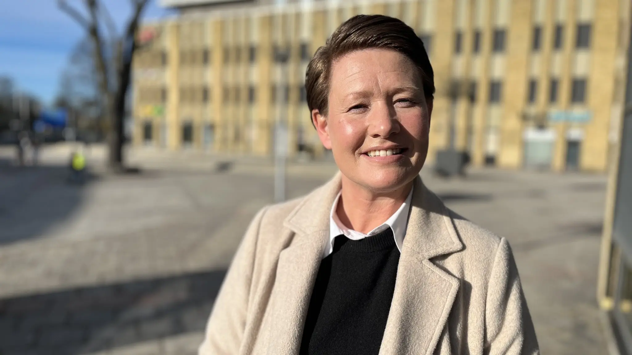 Camilla Freedeke i beige kavaj och svart tröja under, står  i solen på ett torg i Kalmar och ler. hon är korthårig och kisar lite.