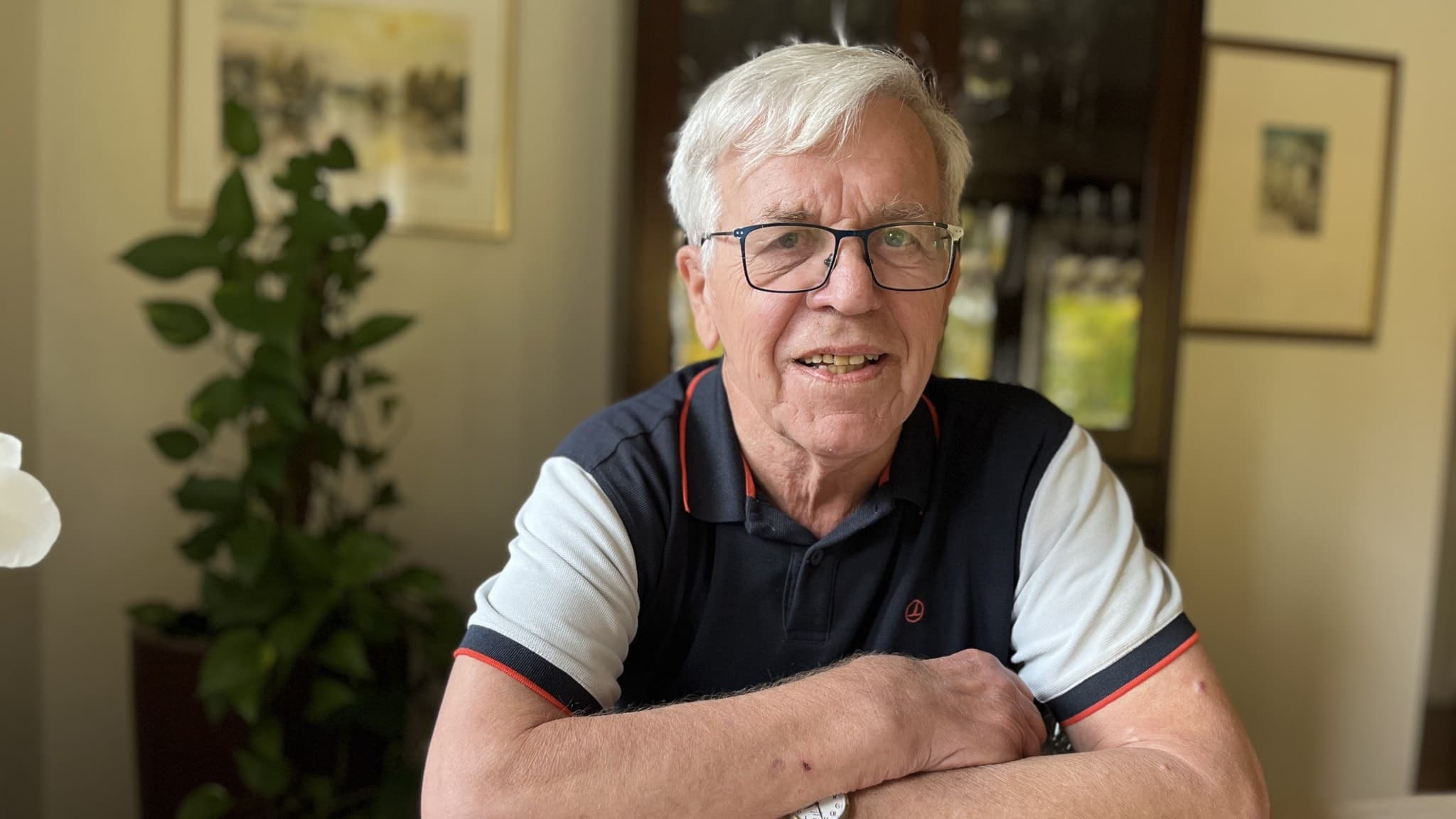 Ingvar Gunnarsson, en äldre man med vitt hår, sitter vid ett bord hemma. Han bär en kortärmad pikétröja och ett armbandsur. Bakgrunden visar växter och ett vitrinskåp i mörkt trä.