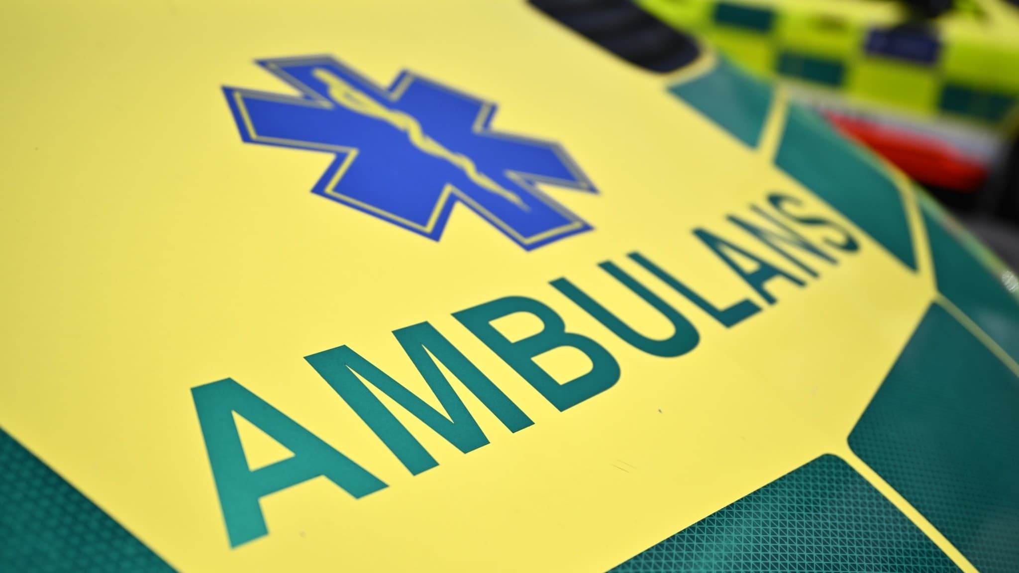 Närbild på en gul och grön ambulans med texten 'AMBULANS' på motorhuven, parkerad i Mariehäll, västra Stockholm, efter rapporter om en misstänkt skottlossning.