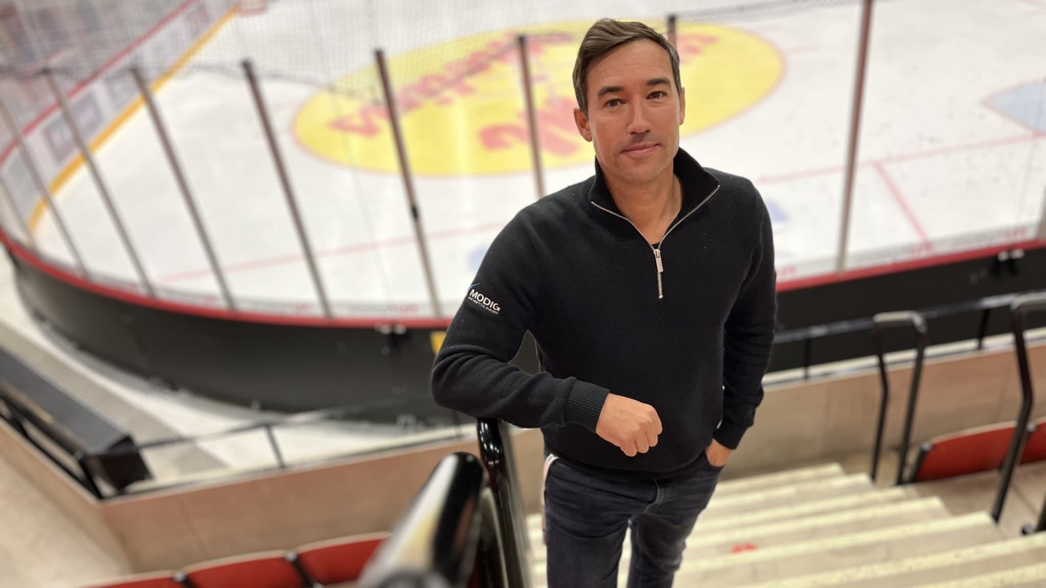 En man står på en trappa i en ishall med en hockeyrink i bakgrunden. Han är klädd i en mörk tröja och jeans. Rinken har reklam på isen och läktarstolar syns runt omkring.