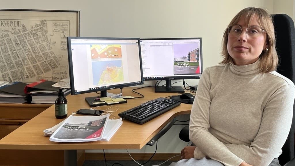 Åsa Bejemar, plansamordnare på Borgholms kommun, sitter vid ett skrivbord med två datorskärmar som visar kartor och planer. På bordet ligger dokument och en flaska syns bredvid.