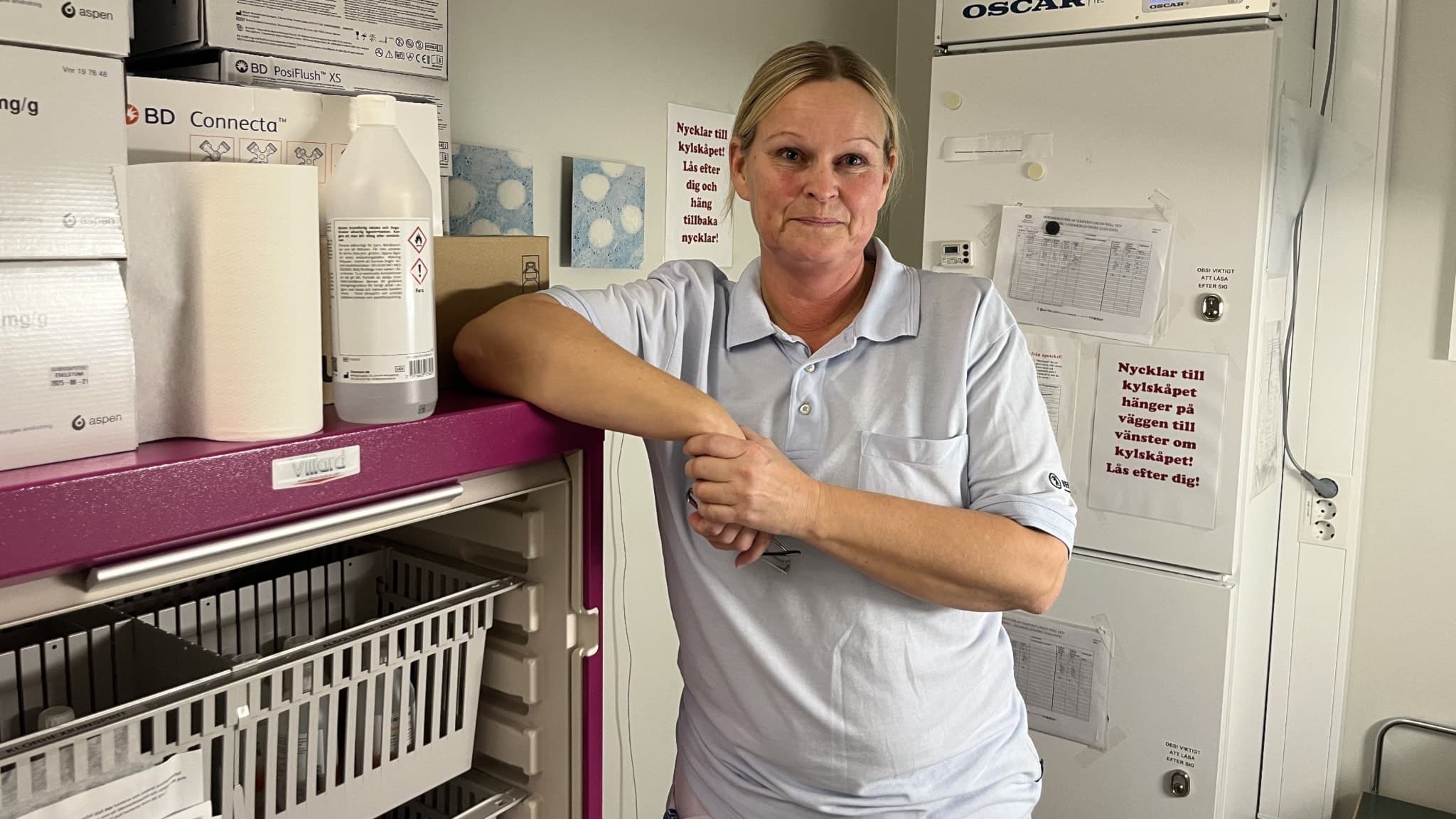 Anne Rydén står inne på Vårdcentralen City, lutad mot en hylla med medicinska förnödenheter. Hon bär vårdkläder och befinner sig i ett rum med vit vägg och medicinsk utrustning.
