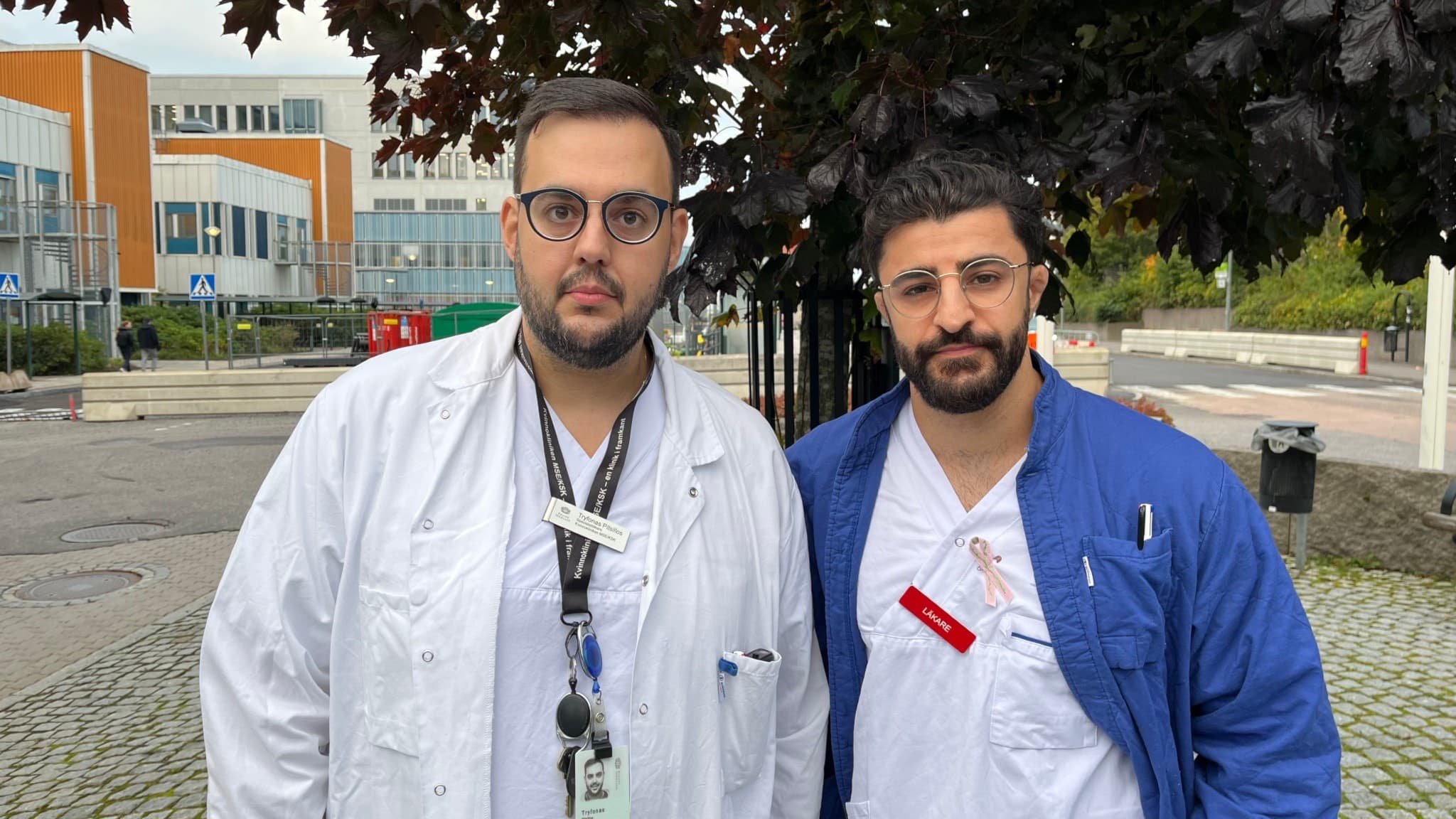 Tryfonos Pitsillos och Dilan Kadir, klädda i sjukhusuniformer, står utomhus framför ett modernt byggnadskomplex med orange och vita detaljer. Trädgrenar syns i förgrunden.