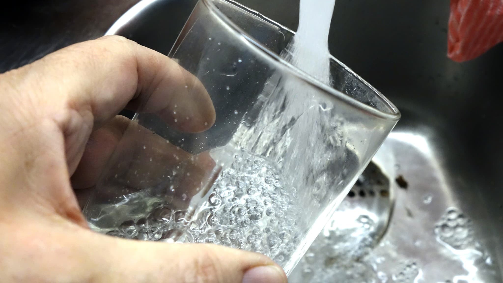 En hand håller ett glas under rinnande vatten i en diskho, med fokus på vattenstrålen och glaset. Bilden symboliserar tillgång till rent dricksvatten.