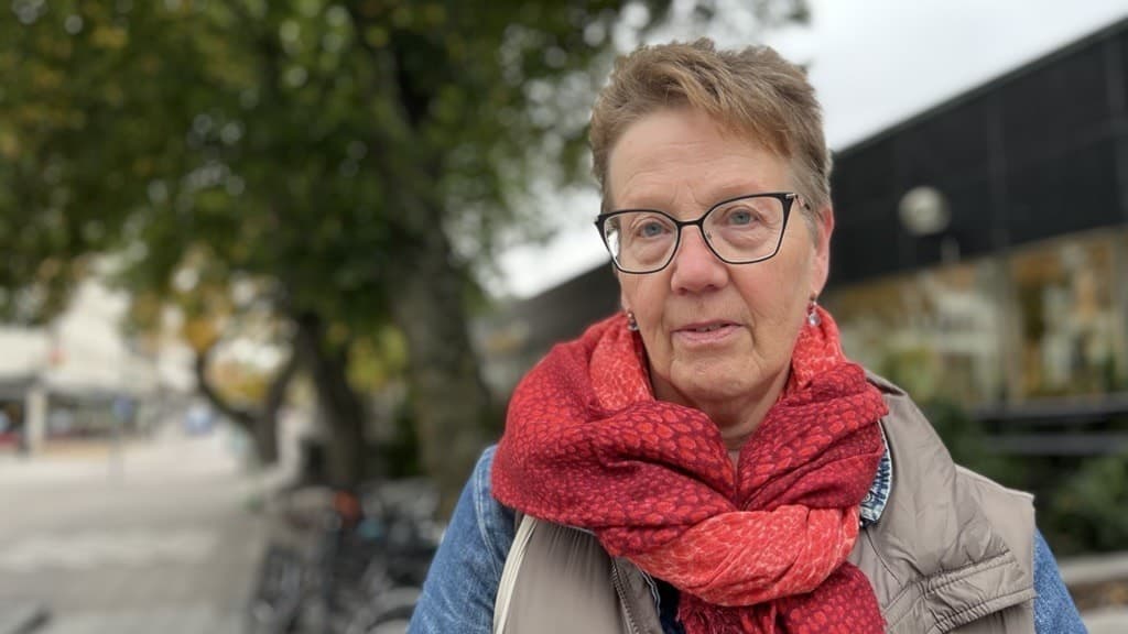 Marja från Eskilstuna står utomhus med en röd halsduk och beige jacka, omgiven av cyklar och träd, och uttrycker svårigheter med att få tid hos läkare.