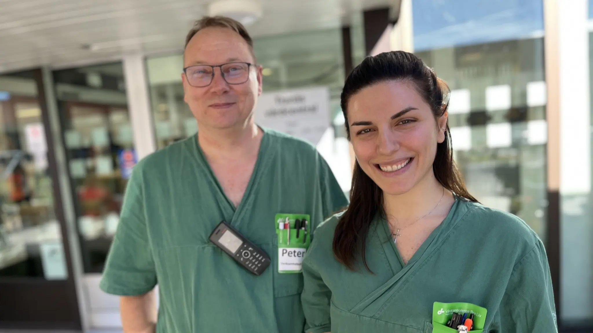 Två sjukvårdspersonal i gröna arbetskläder står framför en byggnad med glasfönster. Peter Lundin Johansson och Daisy Khaled Massoud bär namnbrickor och sjukvårdsutrustning.