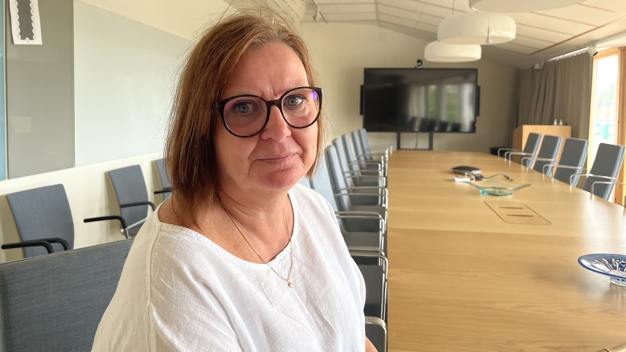 Kvinna i vit blus sitter i ett modernt konferensrum med ett långt bord, omgivna av stolar och en stor skärm på väggen.