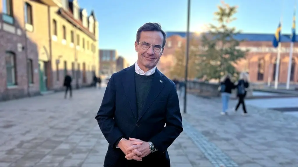 Statsminister Ulf Kristersson står utomhus på en stenlagd gata, klädd i kostym, med händerna knäppta framför sig. Svenska flaggor syns i bakgrunden, omgivningen har inslag av byggnader och träd.