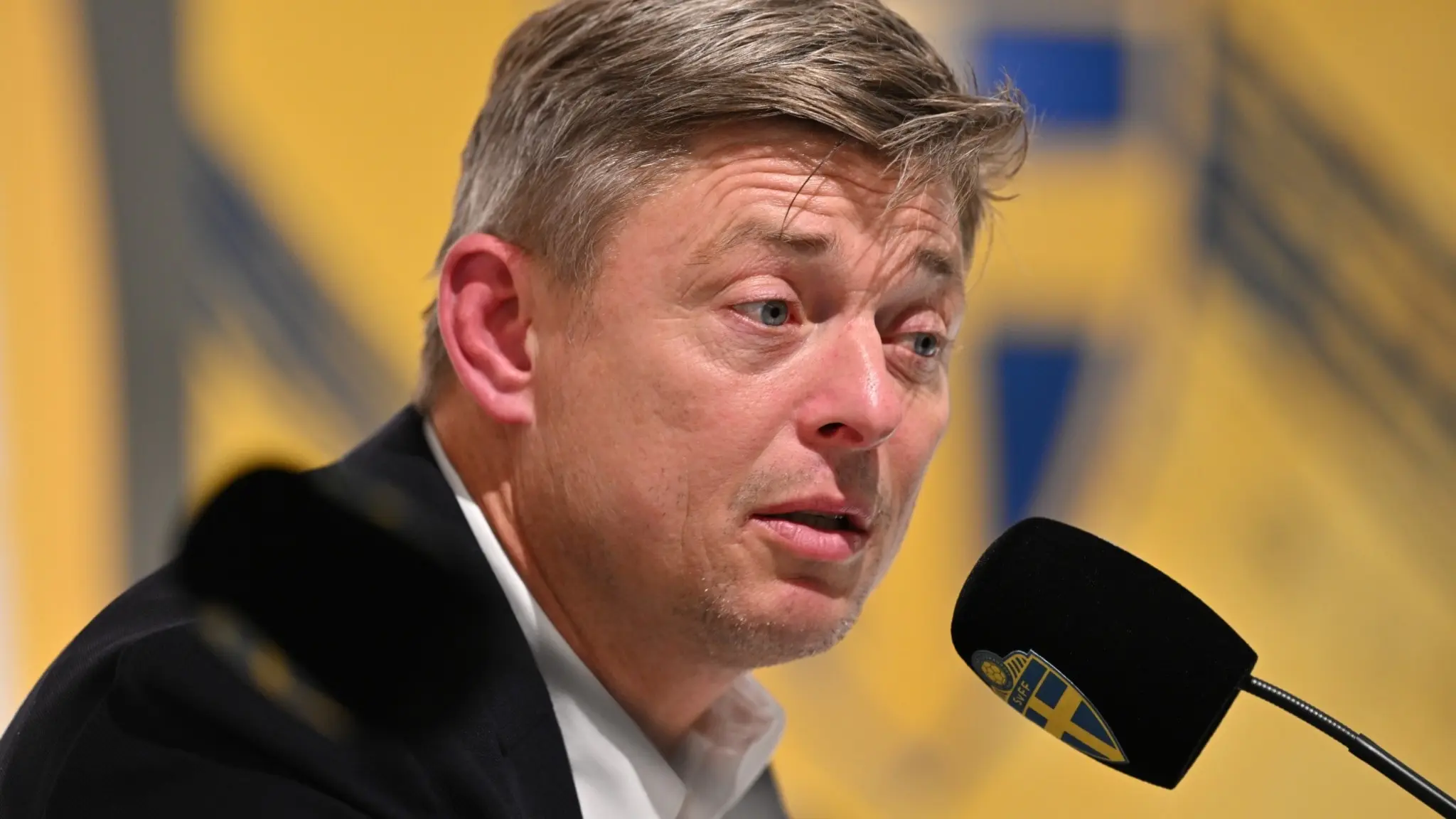 Sveriges förbundskapten Jon Dahl Tomasson talar vid en pressträff efter VM-kvalmatchen i fotboll mellan Sverige och Kosovo på Ullevi, med gul bakgrund och en mikrofon i fokus.