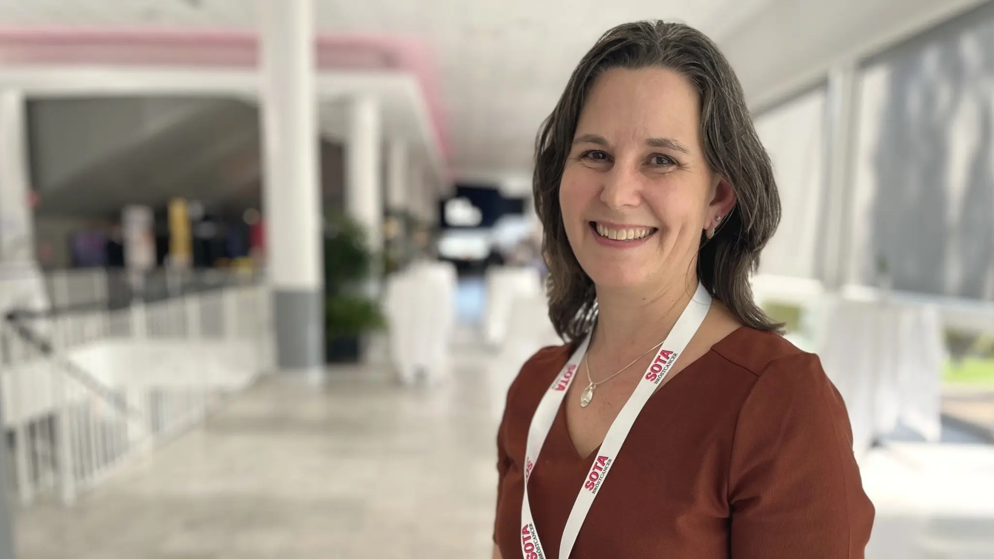 Anna Bodén, patolog på Linköpings Universitetssjukhus, står inomhus i en ljus entrémiljö med vit namnbricka runt halsen. Hon är klädd i en rödbrun tröja och i bakgrunden syns en modern sjukhusmiljö.