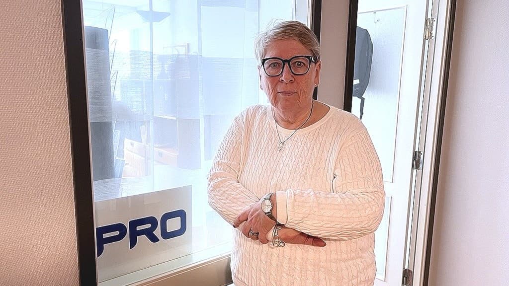 En kvinna med ljus stickad tröja och armarna i kors står inomhus framför ett glasfönster.