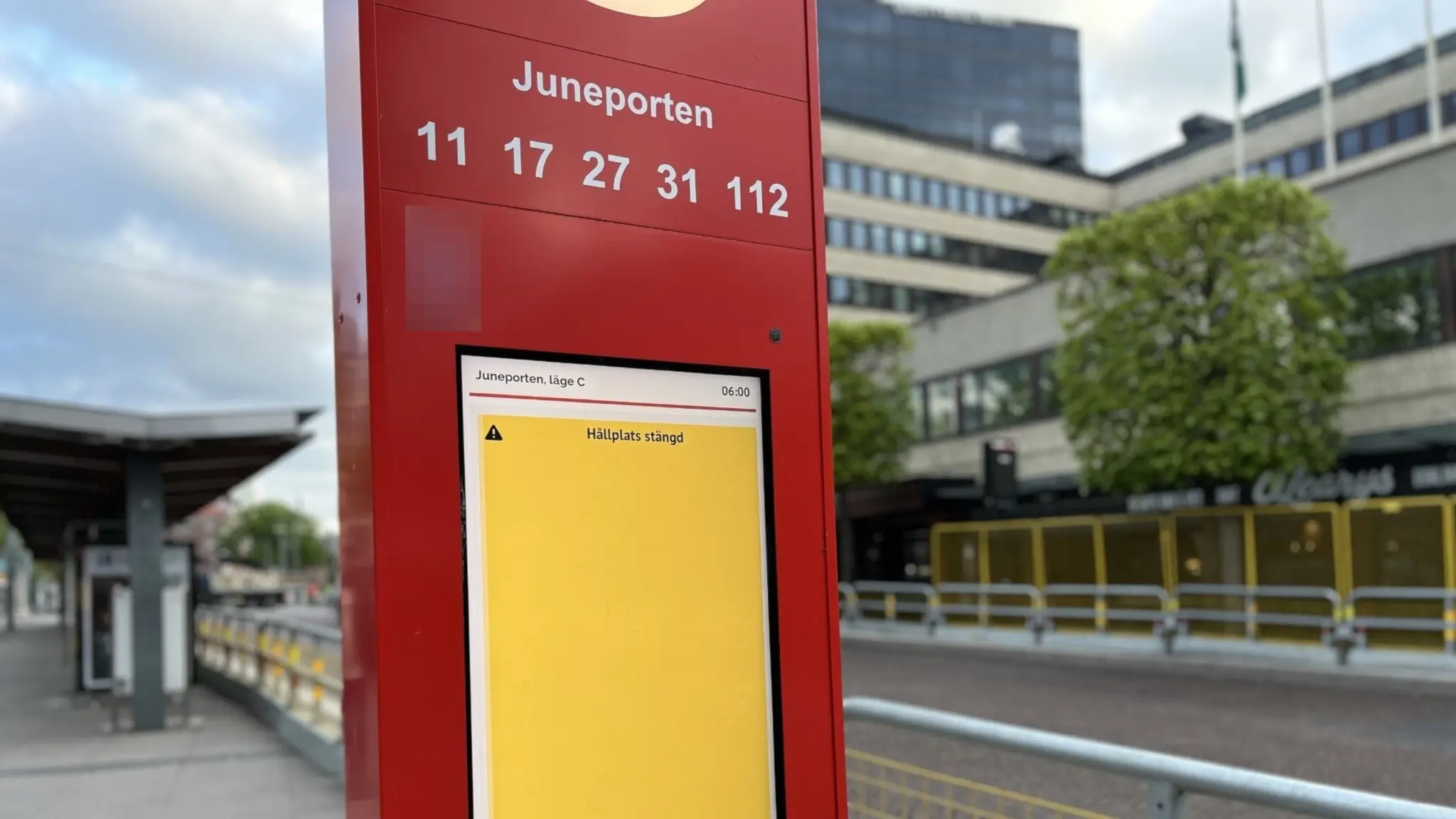 En röd informationspelare vid Juneporten med busshållplats i bakgrunden. Skylten visar linjenummer. På skärmen står det "hållplats avstängd". Ett flervåningshus och grönskande träd syns runt hållplatsen. Molnigt väder.