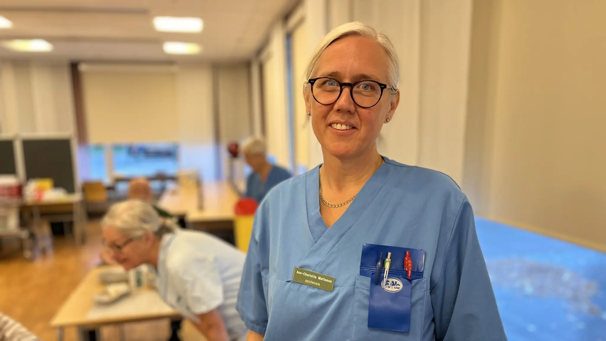 Anna-Charlotte Matsson, klädd i blå sjukhusklädsel, står i en ljus vårdmiljö med stora fönster. I bakgrunden syns andra vårdanställda som utför arbetsuppgifter vid bord.