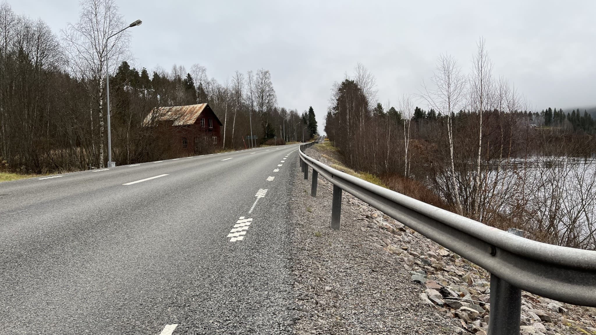 Riksväg 62 slingrar sig genom ett skogsområde med en sjö på ena sidan och ett trähus på andra sidan.