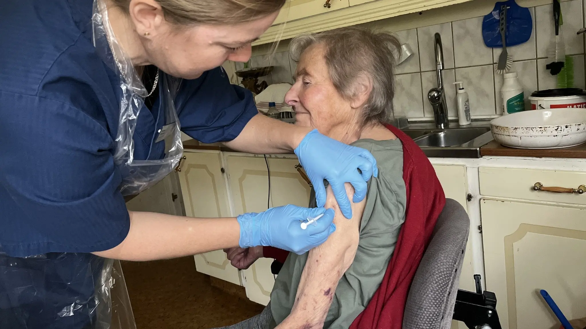 Distriktssjuksköterskan Bettan Nilsson ger covidvaccination till Edith, som sitter i en köksmiljö med en grå tröja och röd kofta. Bettan bär blå skyddshandskar och plastförkläde under vaccineringen.