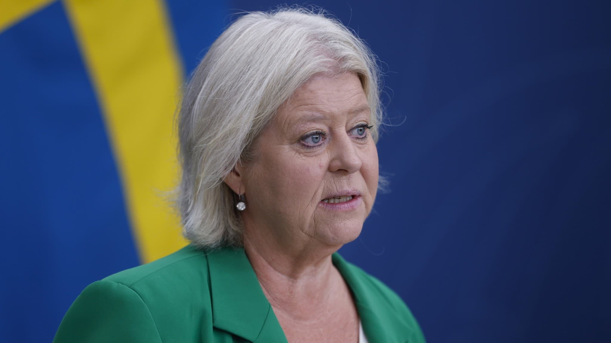 Socialtjänstminister Camilla Waltersson Grönvall (M) talar vid en pressträff. Hon bär en grön kavaj och står framför en svensk flagga i bakgrunden.