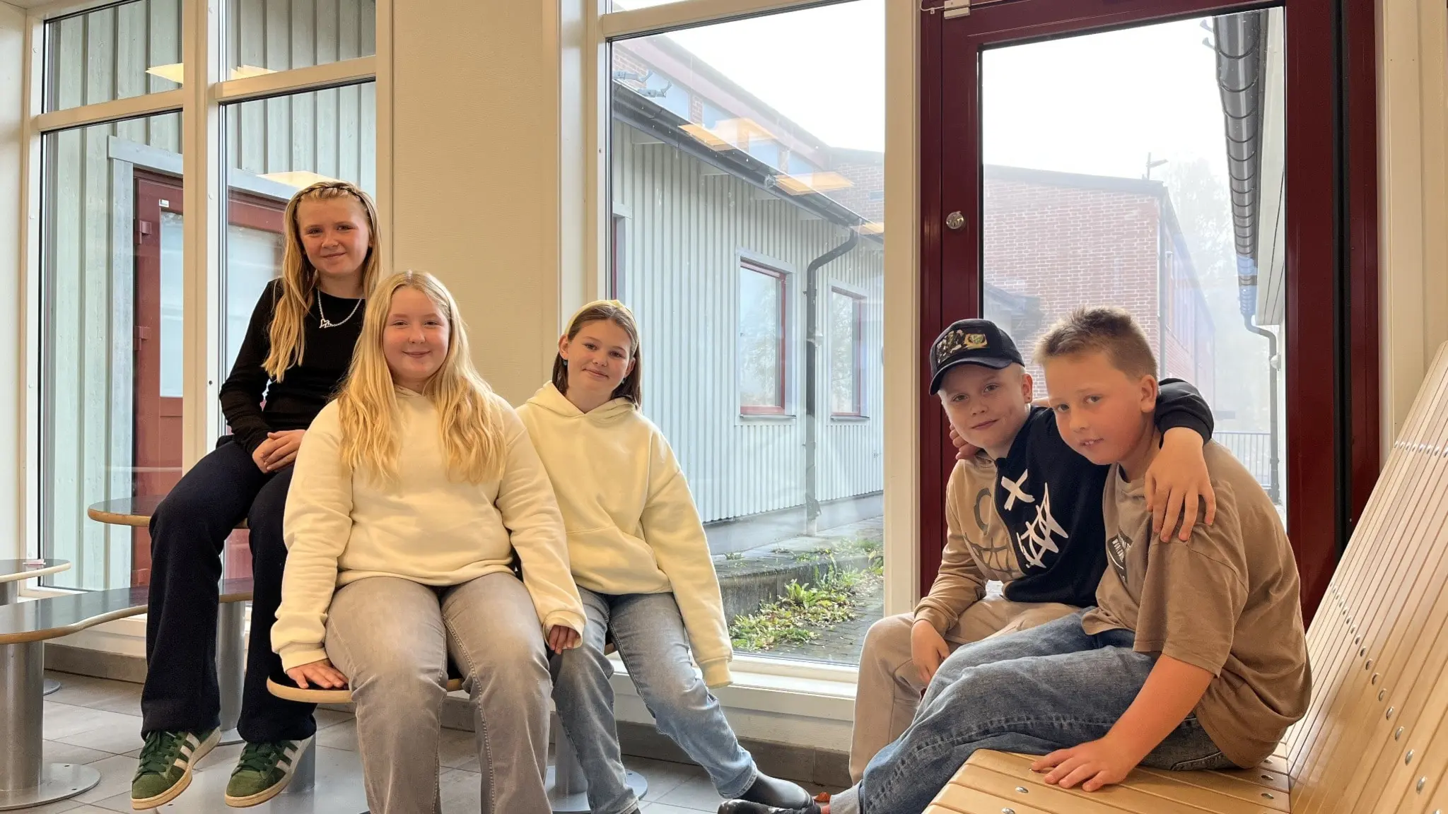 Fem elever från Jättestenskolan i Grums sitter tillsammans vid ett fönster inomhus. De ser avslappnade ut och har på sig vardagskläder. Bakgrunden visar skolbyggnadens utsida.