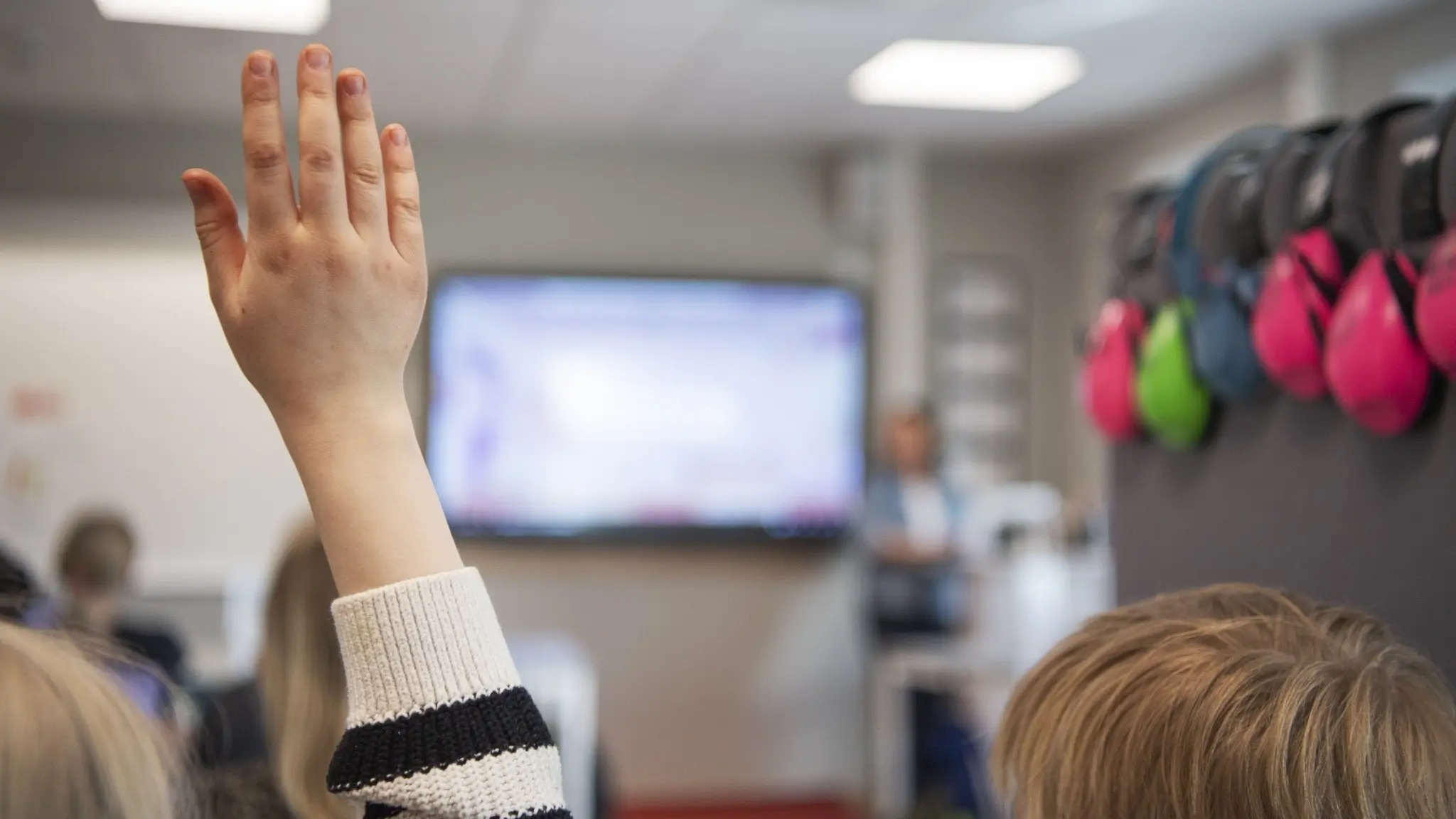 En elev i Västra Götaland räcker upp handen i ett klassrum. I bakgrunden syns en lärare vid en skärm och hörselkåpor i olika färger hängande på väggen. Fokus ligger på elevens hand.