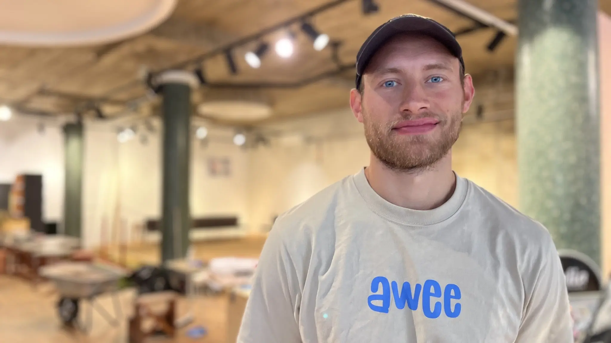 Villiam Dahlström står i en rymlig lokal med industriväggar och taklampor. Han har på sig en ljus t-shirt med texten 'awee'. Lokalen har möbler och arbetsmaterial som tyder på renovering.