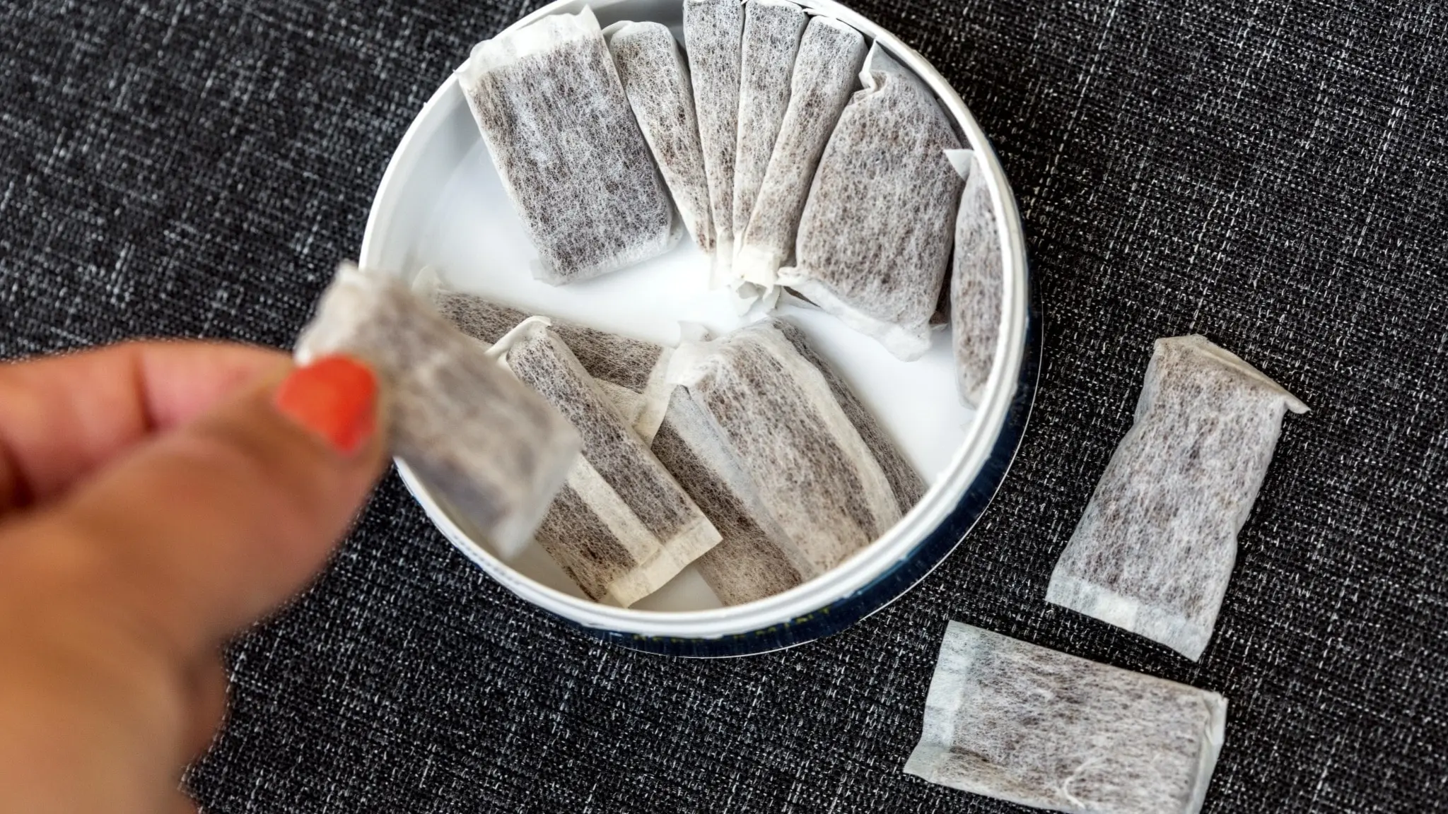 En person håller en portionspåse med snus över en öppen snusburk som ligger på en mörk yta. Inuti burken syns flera snuspåsar.