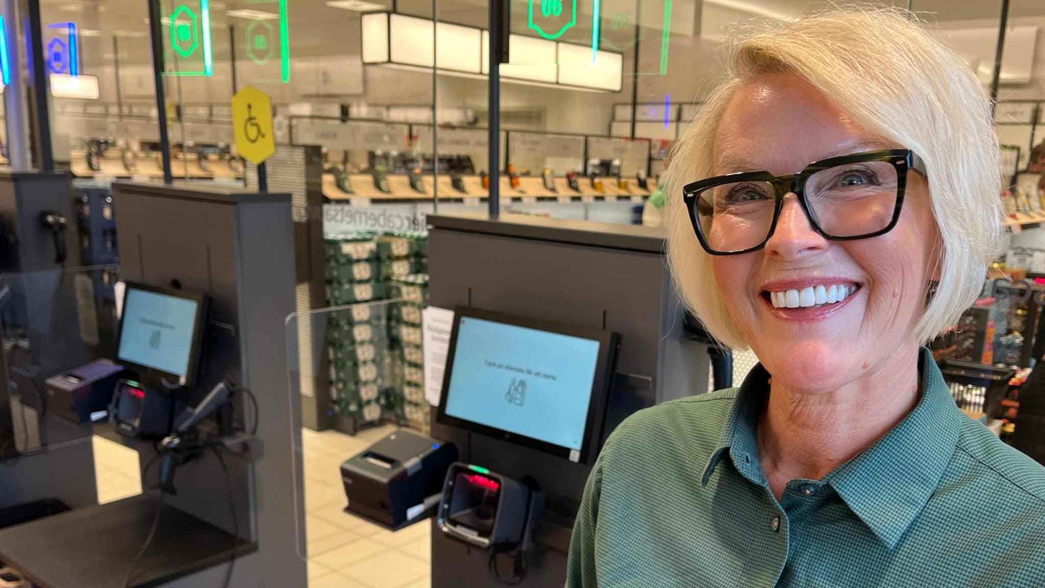 Anna Malmfors från Systembolaget står vid en självbetjäningskassa i en ljus butiksmiljö. I bakgrunden syns hyllor med flaskor och en annan anställd vid en kassa.