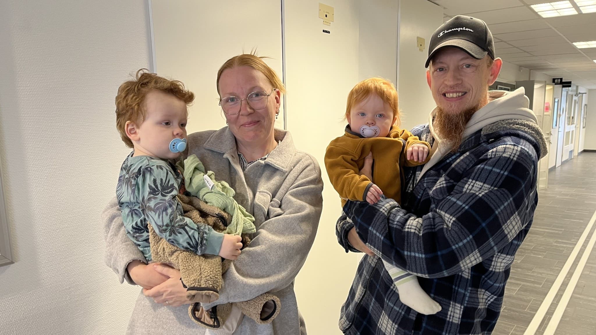 Familjen Saetran står inomhus i en ljus korridor. Två vuxna håller varsitt barn i famnen. 