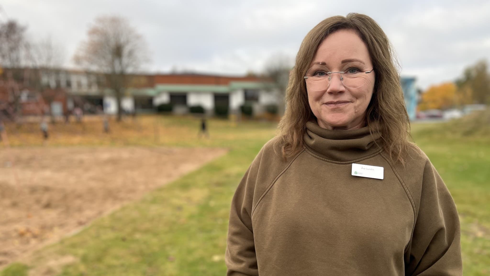 Åsa Lundin, lärare på Tranängskolan i Tranemo, står utomhus på en gräsmatta med skolbyggnader och höstträd i bakgrunden.