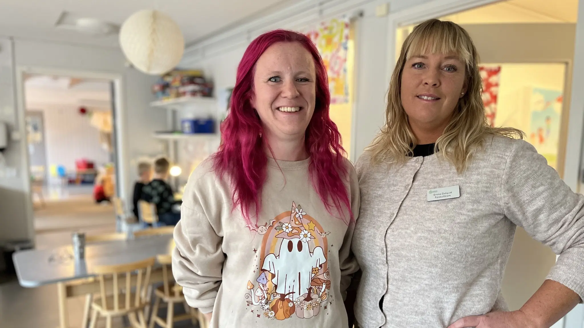 Cecilia Lillhage och Emma Dalquist står i en ljus inomhusmiljö, omgivna av bord och stolar. En pedagogisk miljö med barn i bakgrunden som sitter vid ett bord.