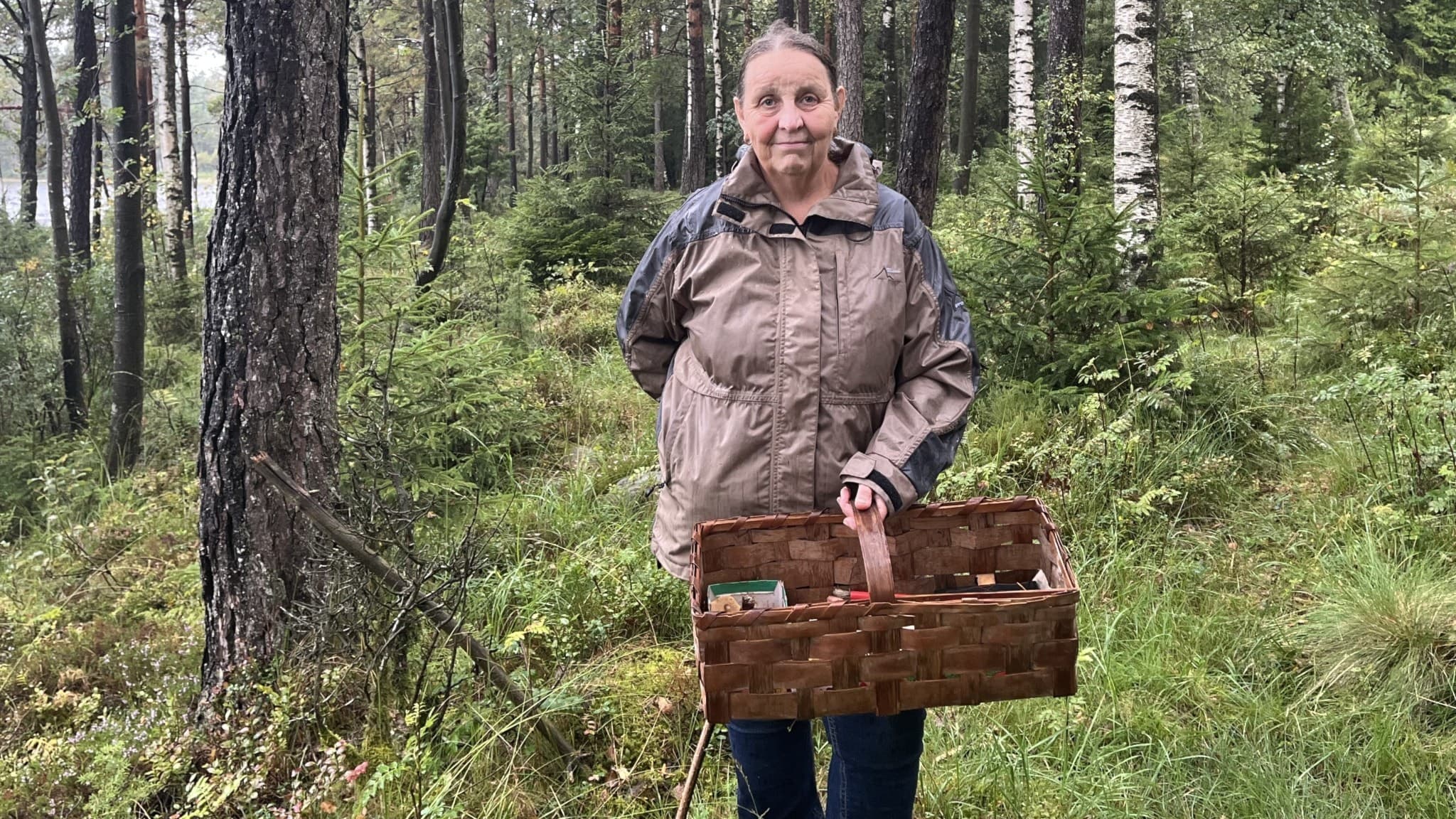 Evy Andreasson, svampkonsulent, står klädd i ljusbrun friluftsjacka i en grönskande skog med en korg fylld av svamp. 