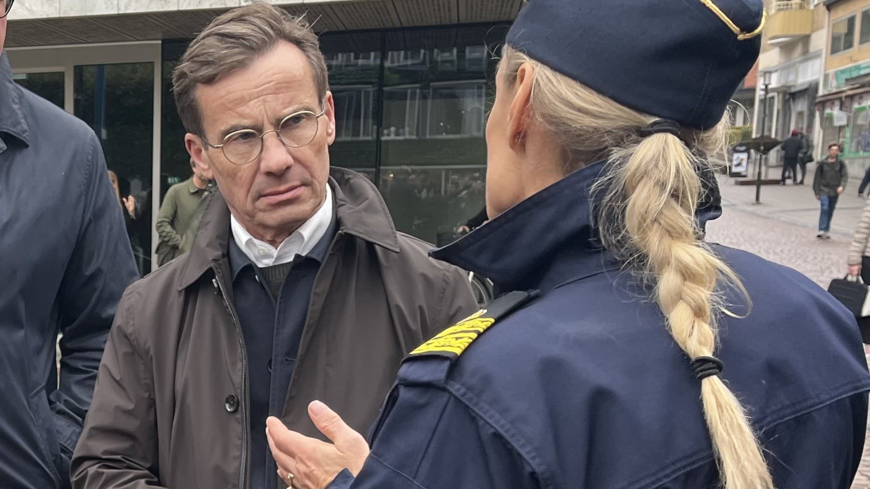 Statsminister Ulf Kristersson samtalar med en polis på en gata i Borås. Han är klädd i mörk kavaj och står i en stadsmiljö med människor och byggnader i bakgrunden.