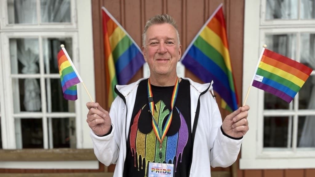Ulf Persson, volontär vid Pride i Ulricehamn, håller två regnbågsflaggor framför en träbyggnad med fler flaggor i bakgrunden. Han bär en Pride-tröja och ett namnkort.