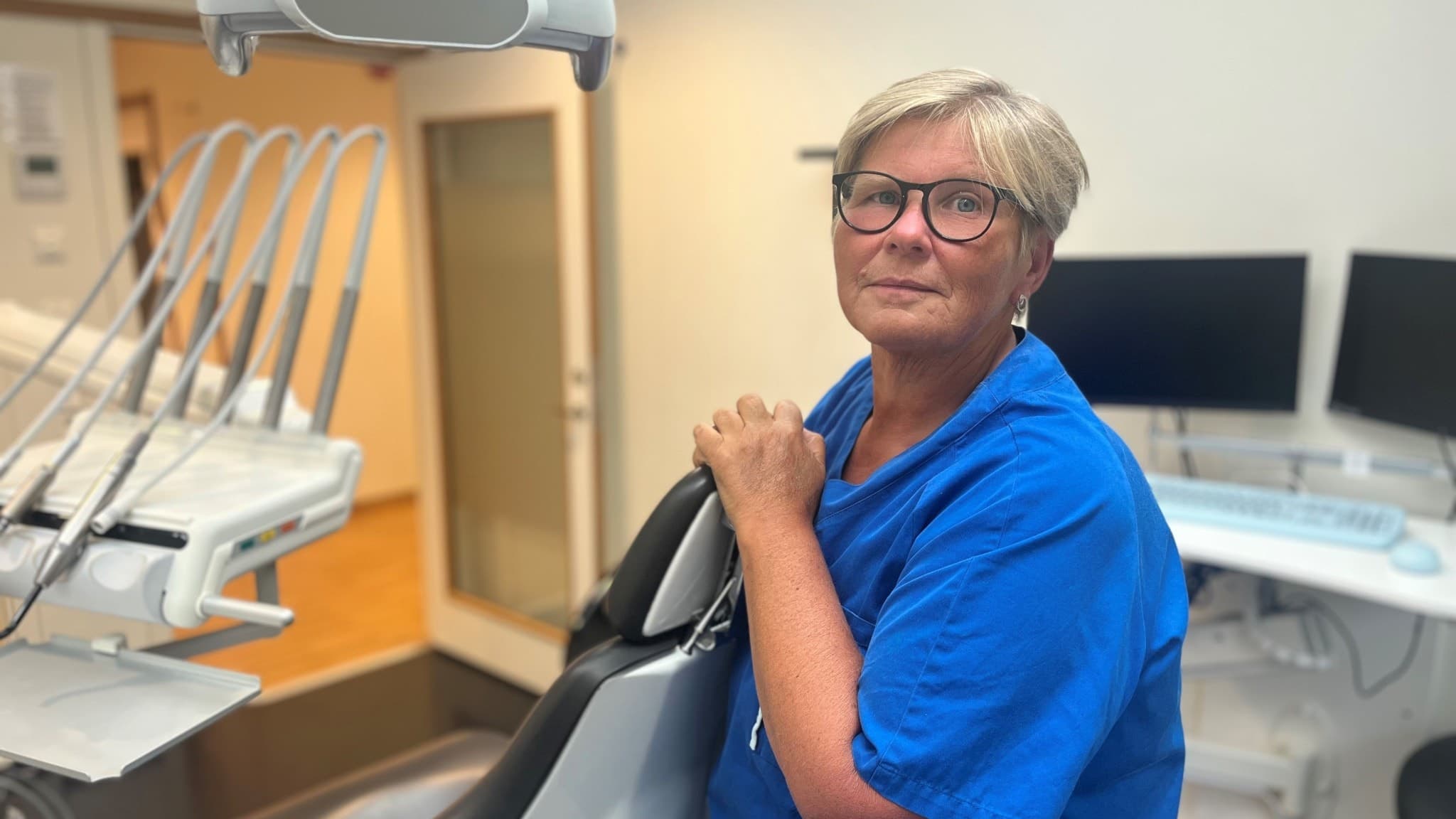 Jaana Sandroos, klinikchef på Folktandvården i Borås, står i en tandläkarmottagning iklädd blå arbetskläder. Tandläkarutrustning och datorer syns i bakgrunden.