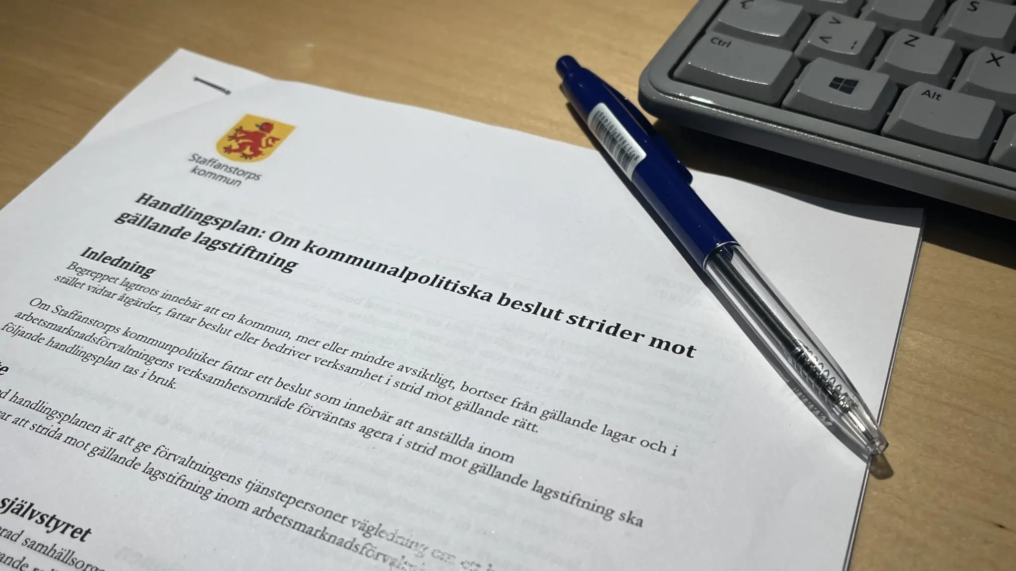 Ett papper med Staffanstorps kommuns handlingsplan om kommunpolitiska beslut strider mot lag. Dokumentet ligger på ett träfärgat skrivbord och en kulspetspenna ligger ovanpå. Uppe till höger syns ett tangentbord. Foto: Erik Adell/Sveriges Radio. 