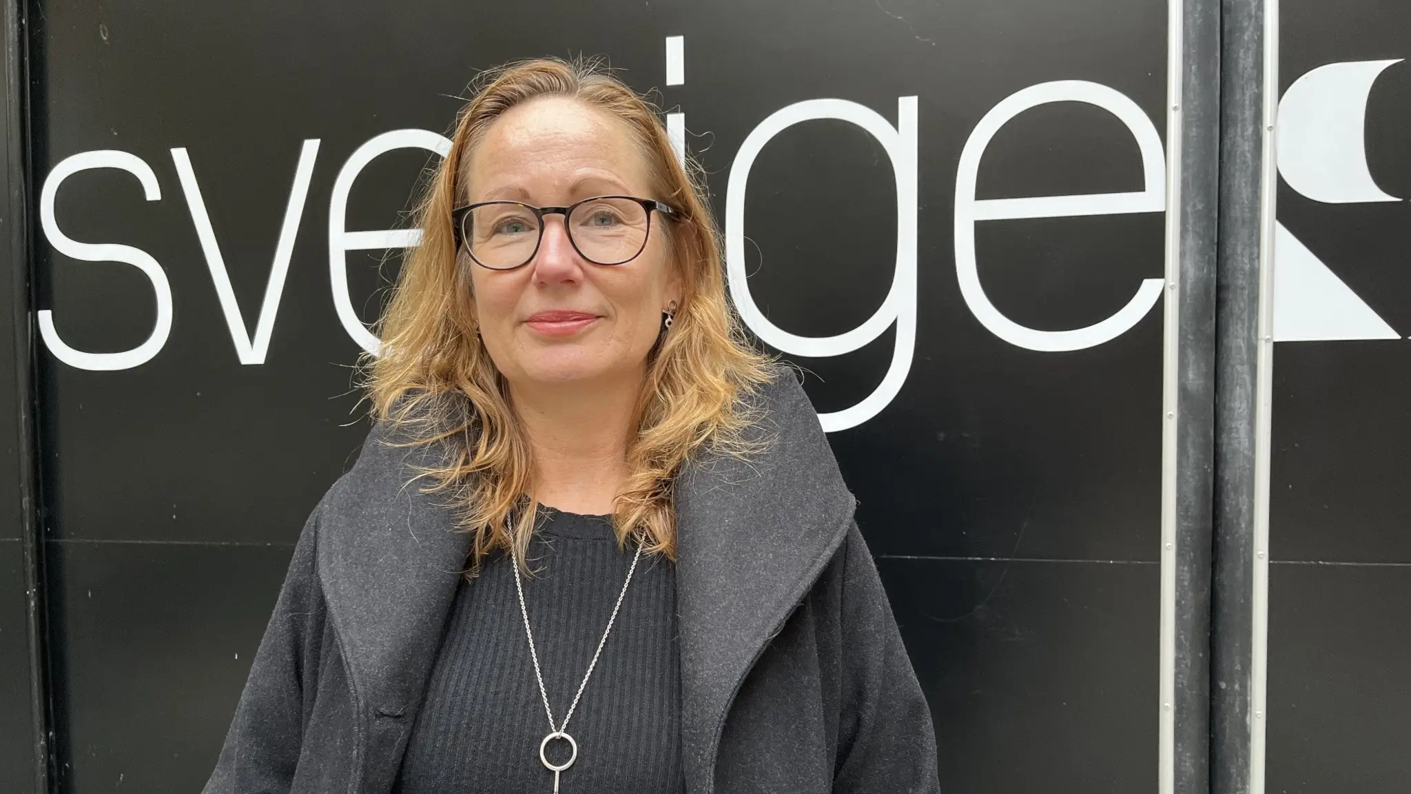 Sara Wettergren (L), ordförande i grundskolenämnden i Malmö, står framför en svart vägg med texten 'Sveriges Radio' synlig i bakgrunden. Hon är klädd i en mörk kappa och har ett silverhalsband.