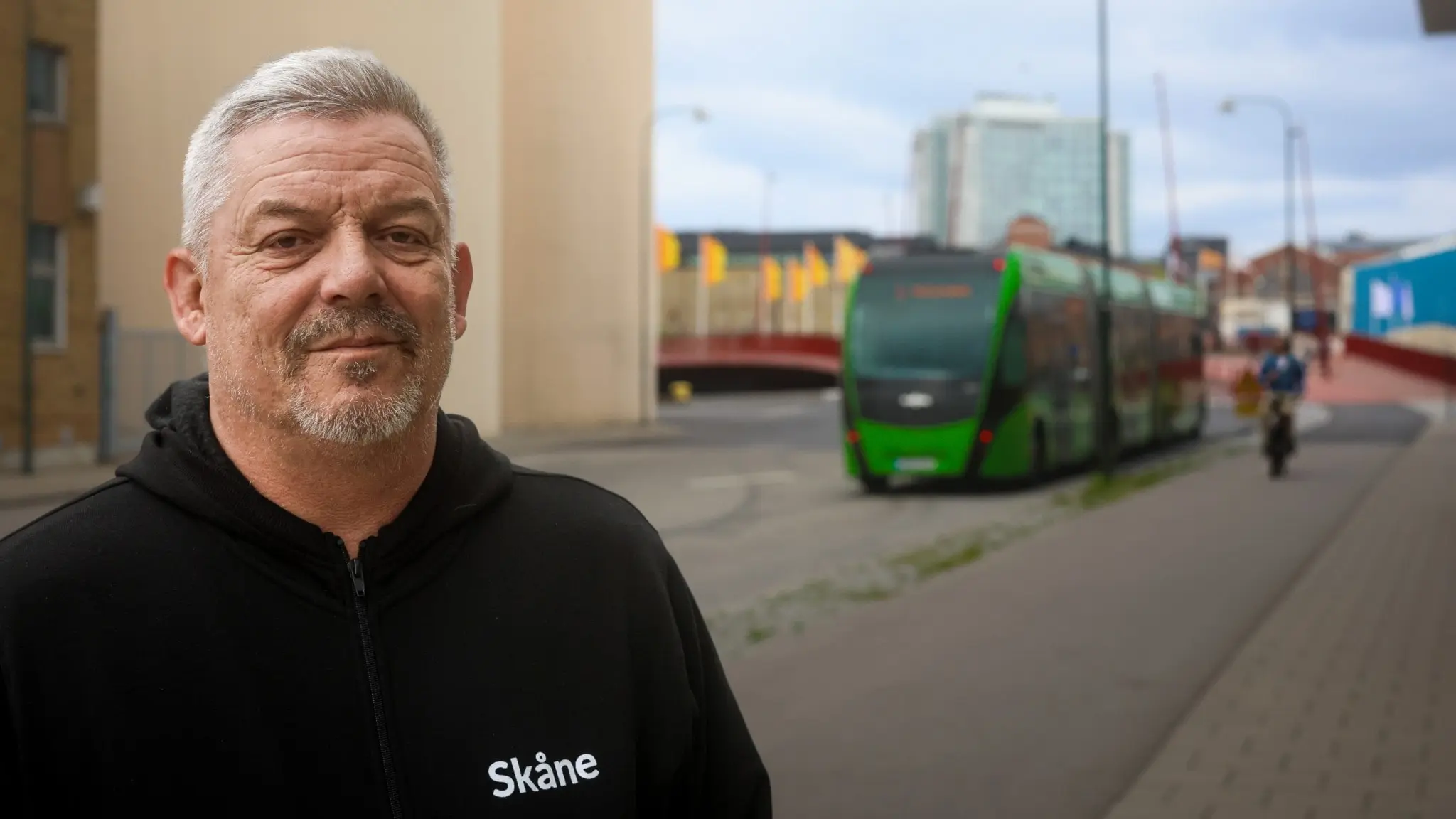 Mikael Olsson, regionalt skyddsombud på Kommunal, står vid Styrmansbron i Malmö med en grön lokaltrafikbuss som kör på vägen bakom honom. 