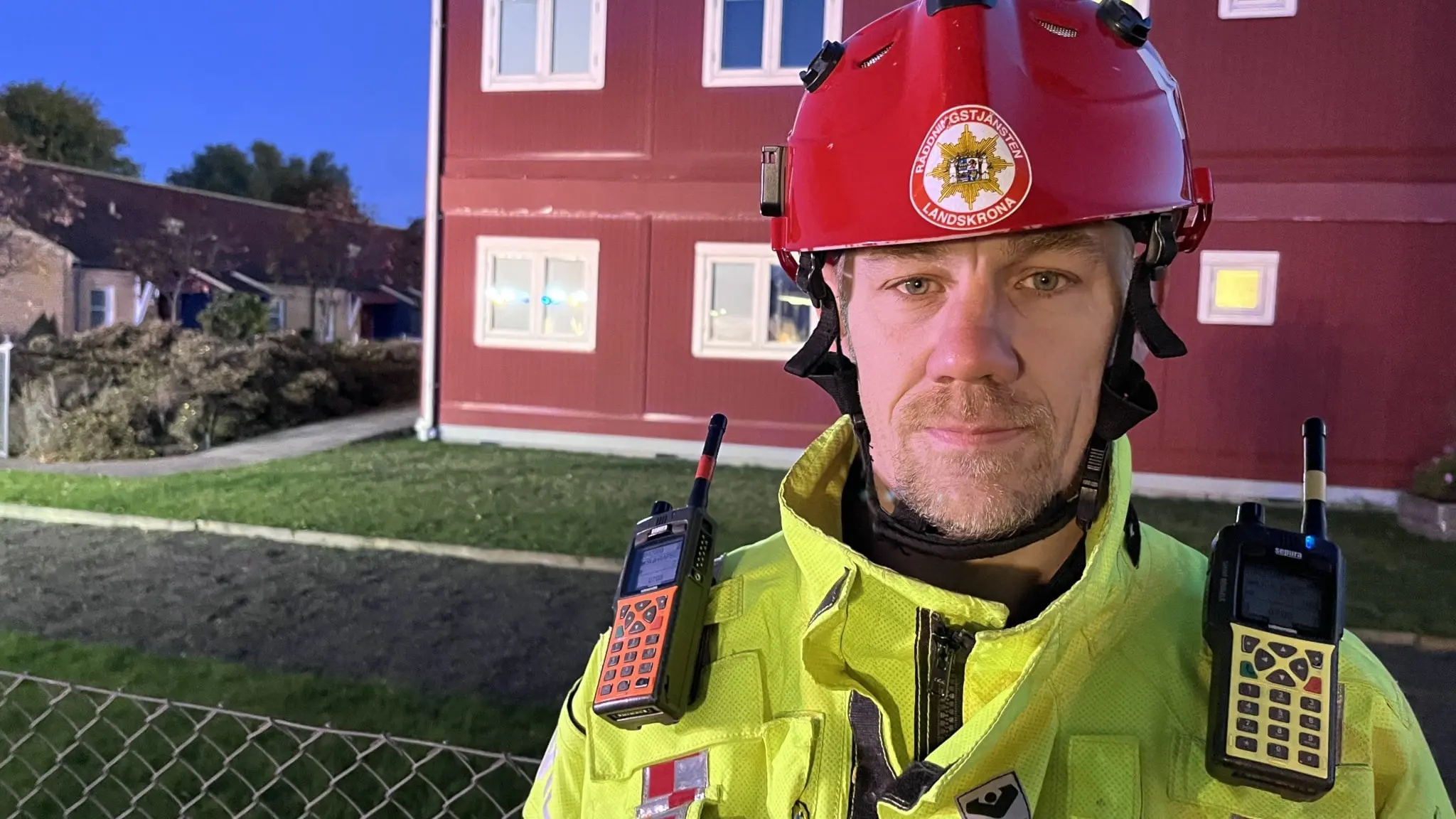 Mattias Lassén, insatsledare vid Räddningstjänsten i Landskrona, står i gul räddningsuniform och röd hjälm framför ett bostadshus, med kommunikationsutrustning på uniformen.