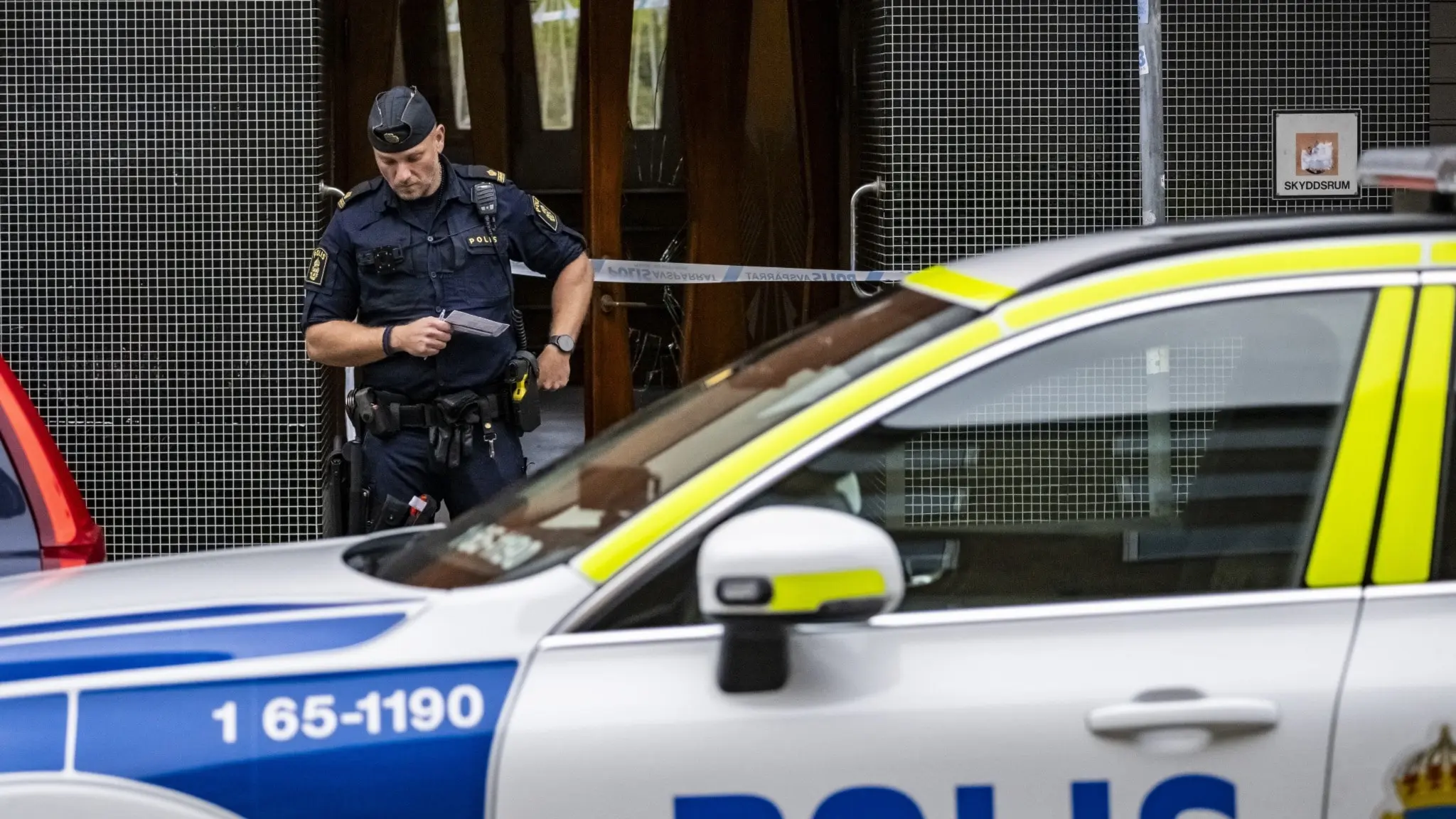 En polis står bakom ett avspärrningsband vid en byggnad i Malmö efter ett misstänkt mordförsök. En polisbil står parkerad framför byggnaden. Händelsen utreds och området är avspärrat.