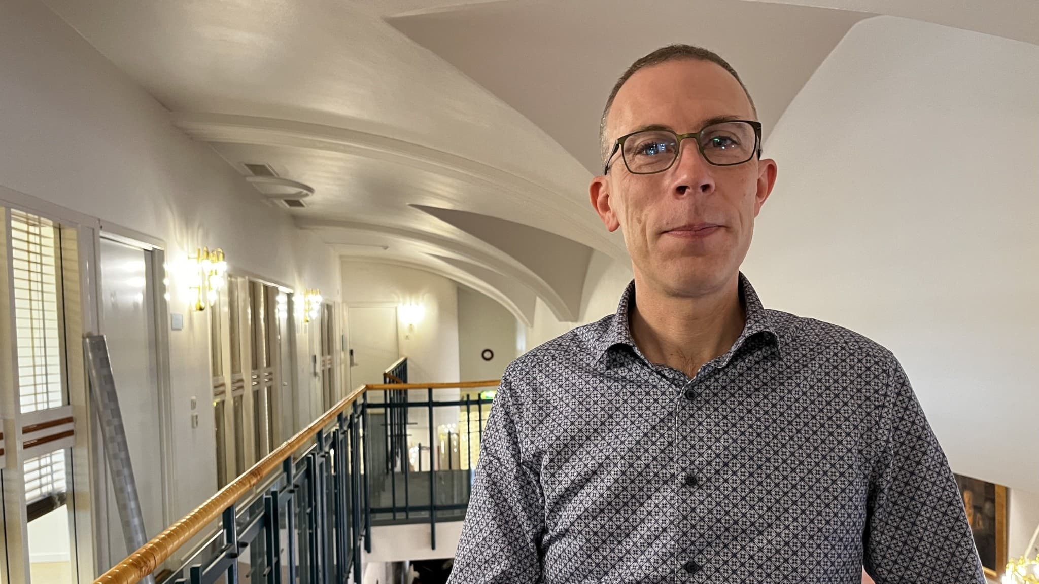 Vilhelm Persson, professor i offentlig rätt, står inomhus vid en gång med välvda tak och glasräcken på Juridicum i Lund. Han bär en mönstrad skjorta. Foto: Anna Bubenko/Sveriges Radio. 