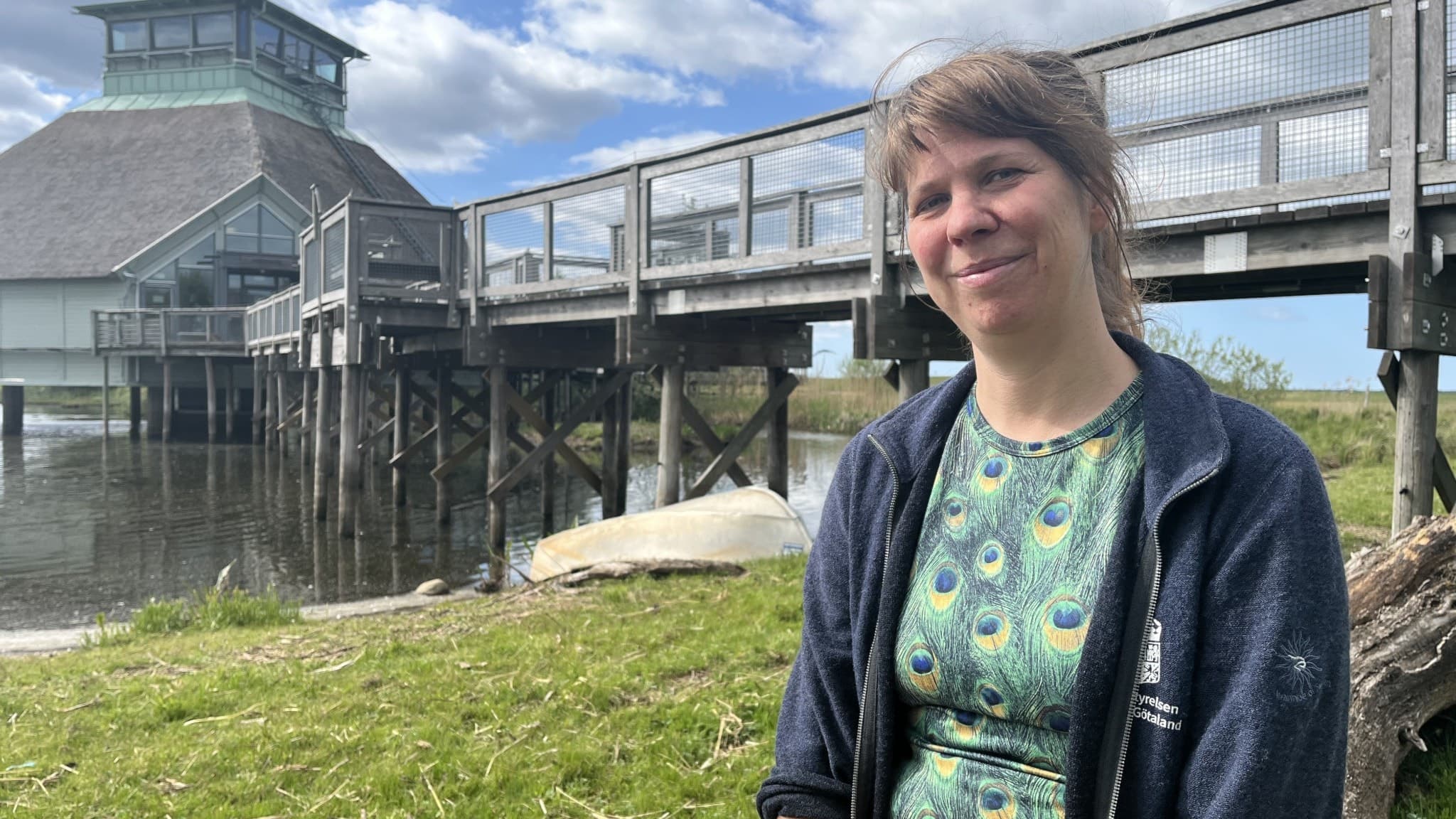 Sofie Stålhand, platschef vid Hornborgasjön, sitter på en gräsplätt med träbyggnaden och bryggan vid naturreservatet i bakgrunden. Himlen är delvis molnig och sjön syns tydligt.