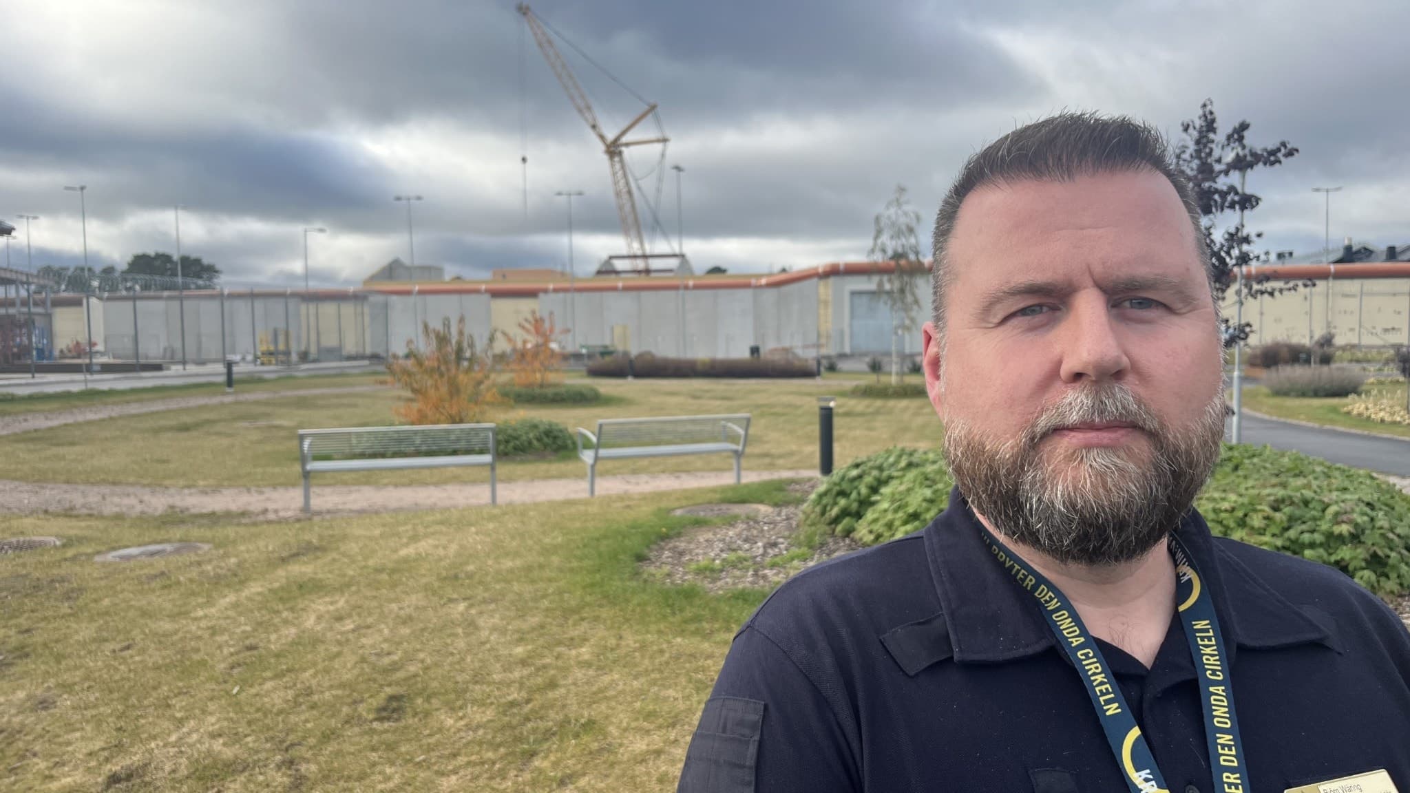 Björn Wäring står utomhus nära en gräsmatta med bänkar och buskar. I bakgrunden syns en byggnad och en lyftkran under molnig himmel.