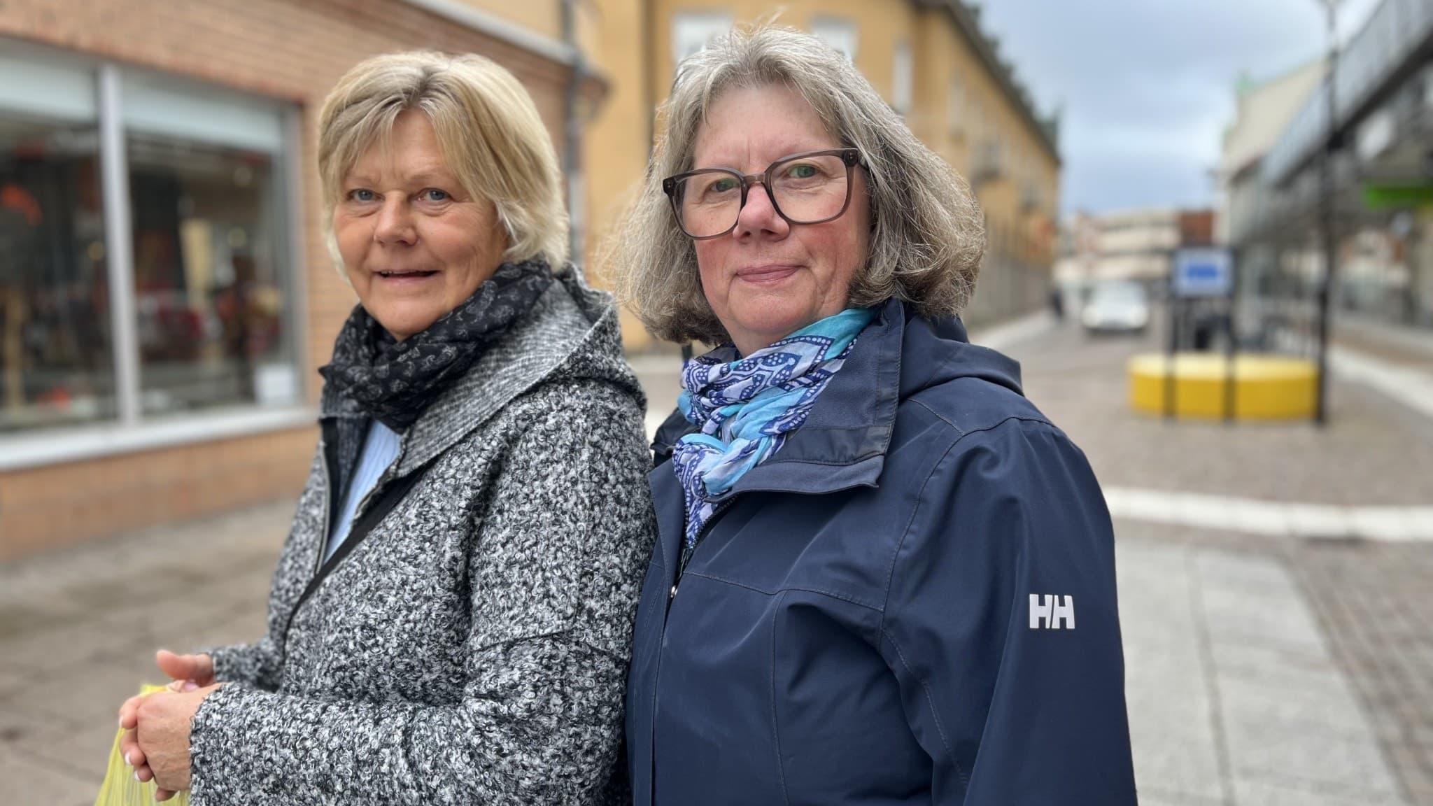 Två kvinnor i jackor står på en gata med butiksfönster och en grå himmel.