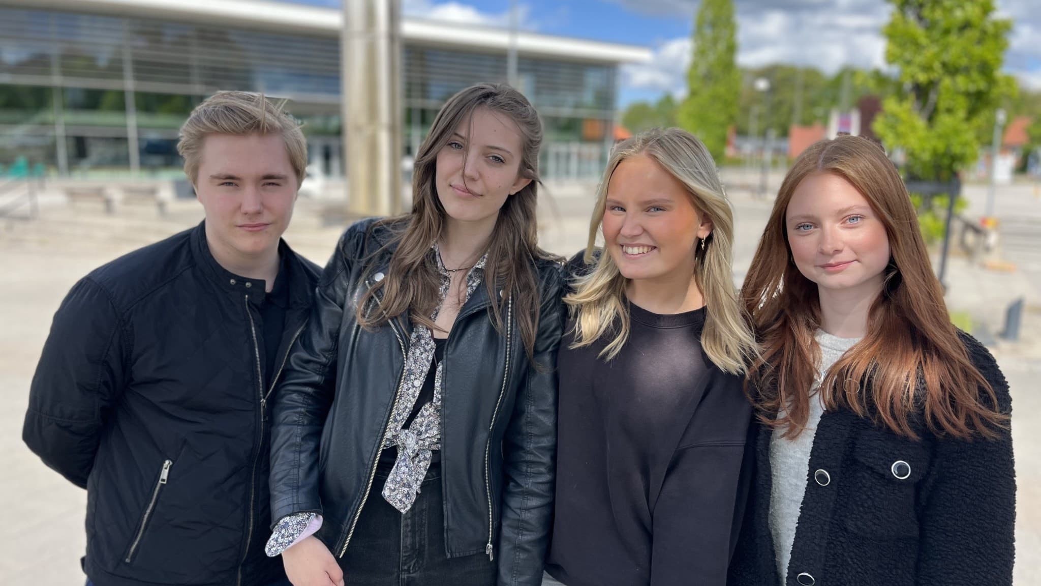 Fyra unga personer, Linus, Signe, Maja och Elsa, står utomhus framför en modern byggnad med glasfasad och grönska i bakgrunden. De är klädda i mörka och neutrala färger.