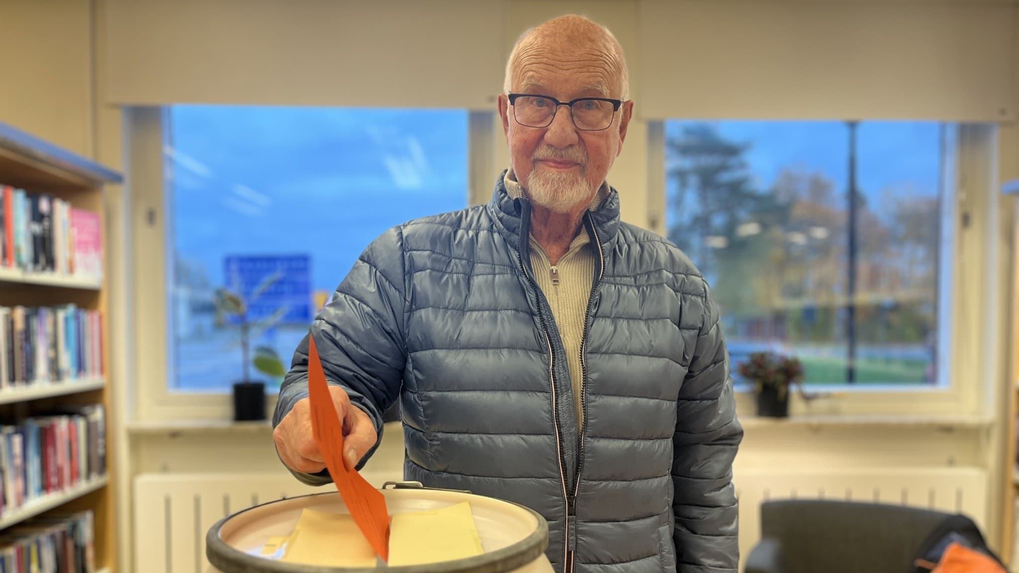 En person klädd i blå dunjacka lägger ett kuvert i en valurna. Bakgrunden visar en hylla med böcker och ett fönster med utsikt över ett grönområde.