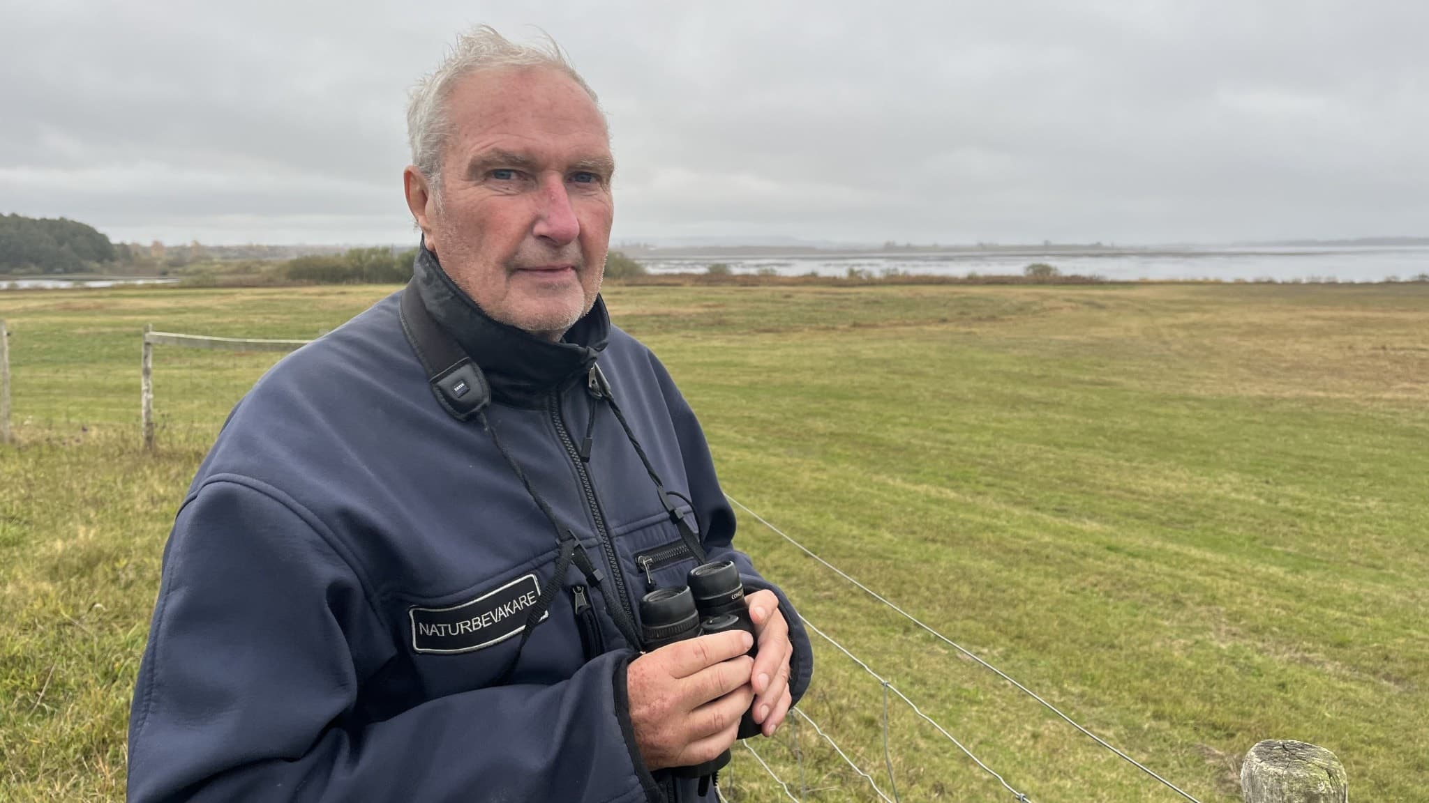 Peder Hedberg Fält, naturvårdshandläggare på Länsstyrelsen, står på en grön äng med en kikare i händerna. I bakgrunden syns ett landskap med vatten och molnig himmel.