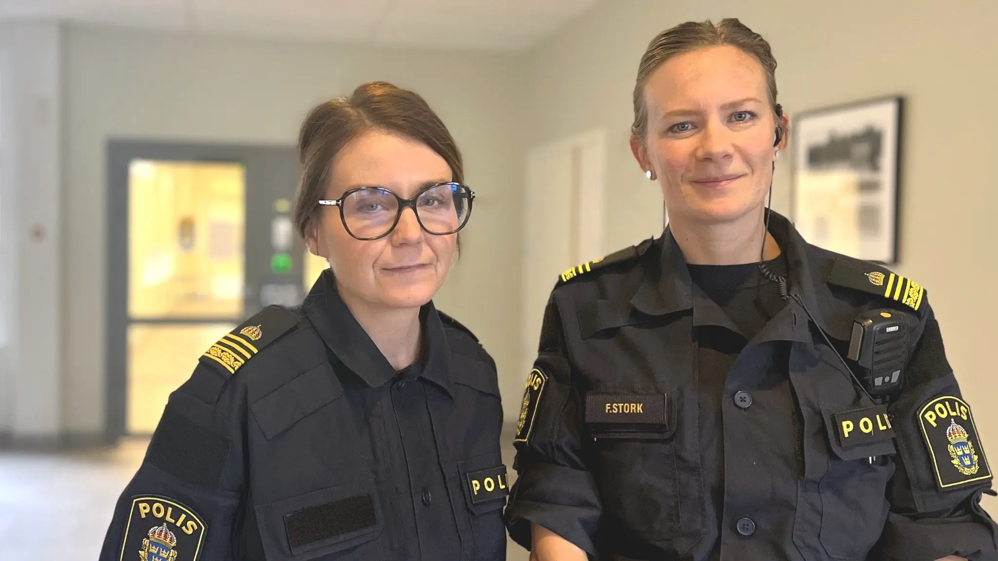 Två poliskvinnor klädda i uniform står i en korridor och tittar in i kameran.