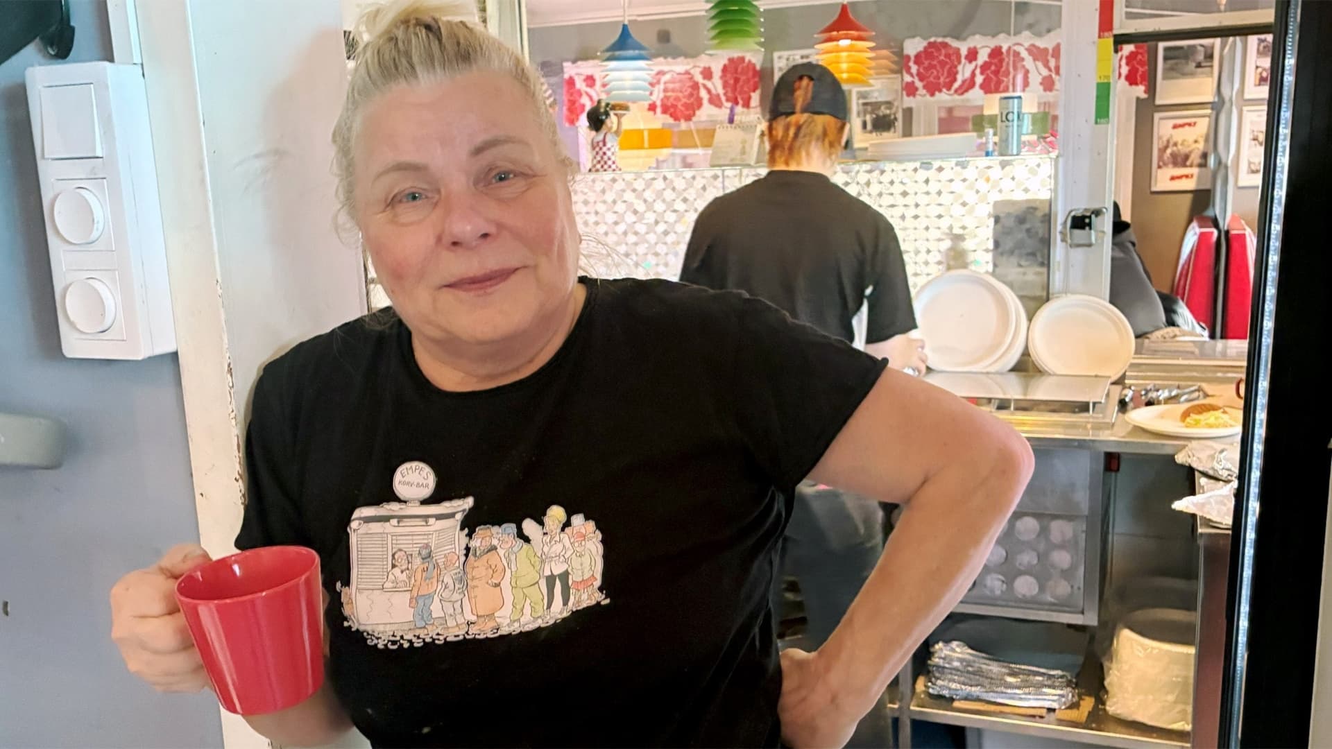 Mona Carlsson, ägare av Empes gatukök, håller en röd kopp i handen. I bakgrunden syns ett kök med färgglada detaljer och en person som arbetar i gatuköket.