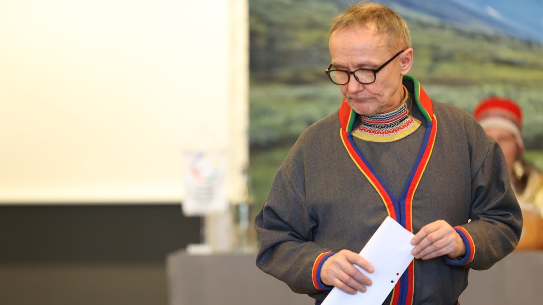 Håkan Jonsson, klädd i sydsamisk kolt, vänder blicken snett neråt medan han håller ett dokument i händerna under ett möte med sametingsdelegater. Bakgrunden visar ett bord och en bild av fjällandskap.