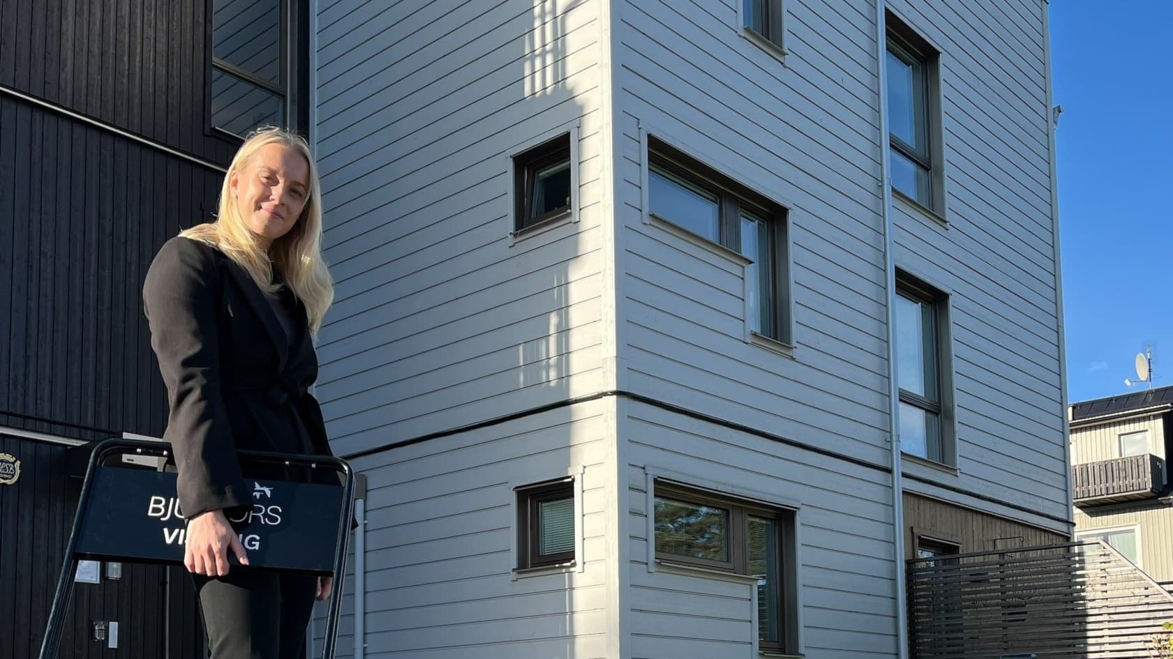 Elvira Henriksson står framför en modern byggnad med vita fasadplattor, klädd i svart och med en laptop i handen. Soligt väder med tydliga skuggor på marken.