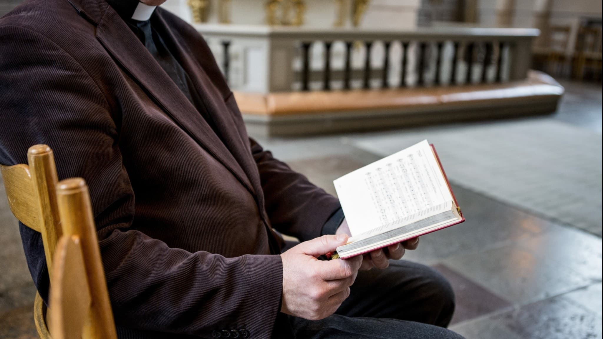 En präst sitter i en kyrkobänk och läser en psalmbok. I bakgrunden syns kyrkans altare och stenbelagd golvyta, vilket förstärker den sakrala atmosfären.